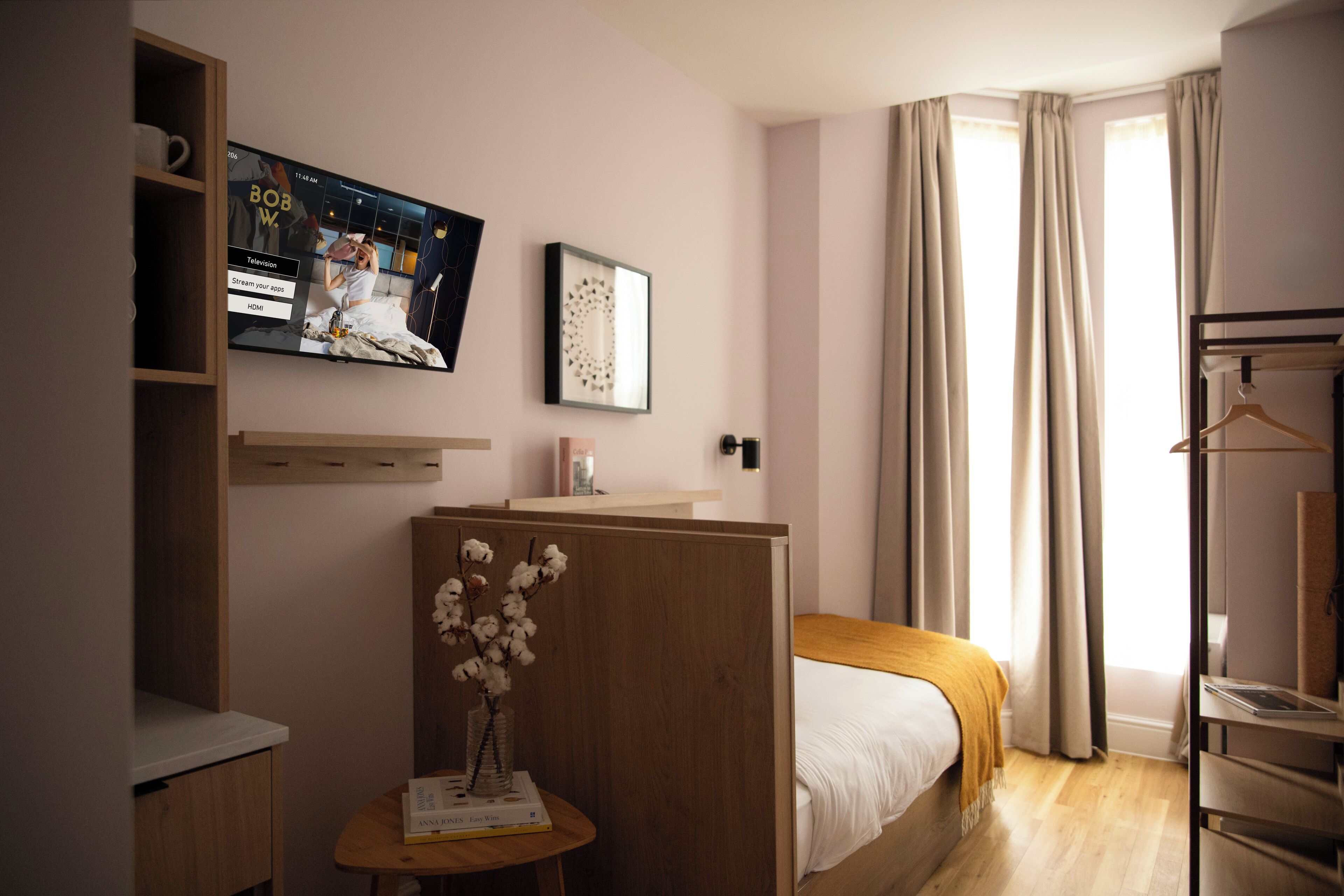 Une chambre d’hôtel avec un lit, un téléviseur fixé au mur, un bureau en bois orné d’un vase de fleurs et un porte-manteau.