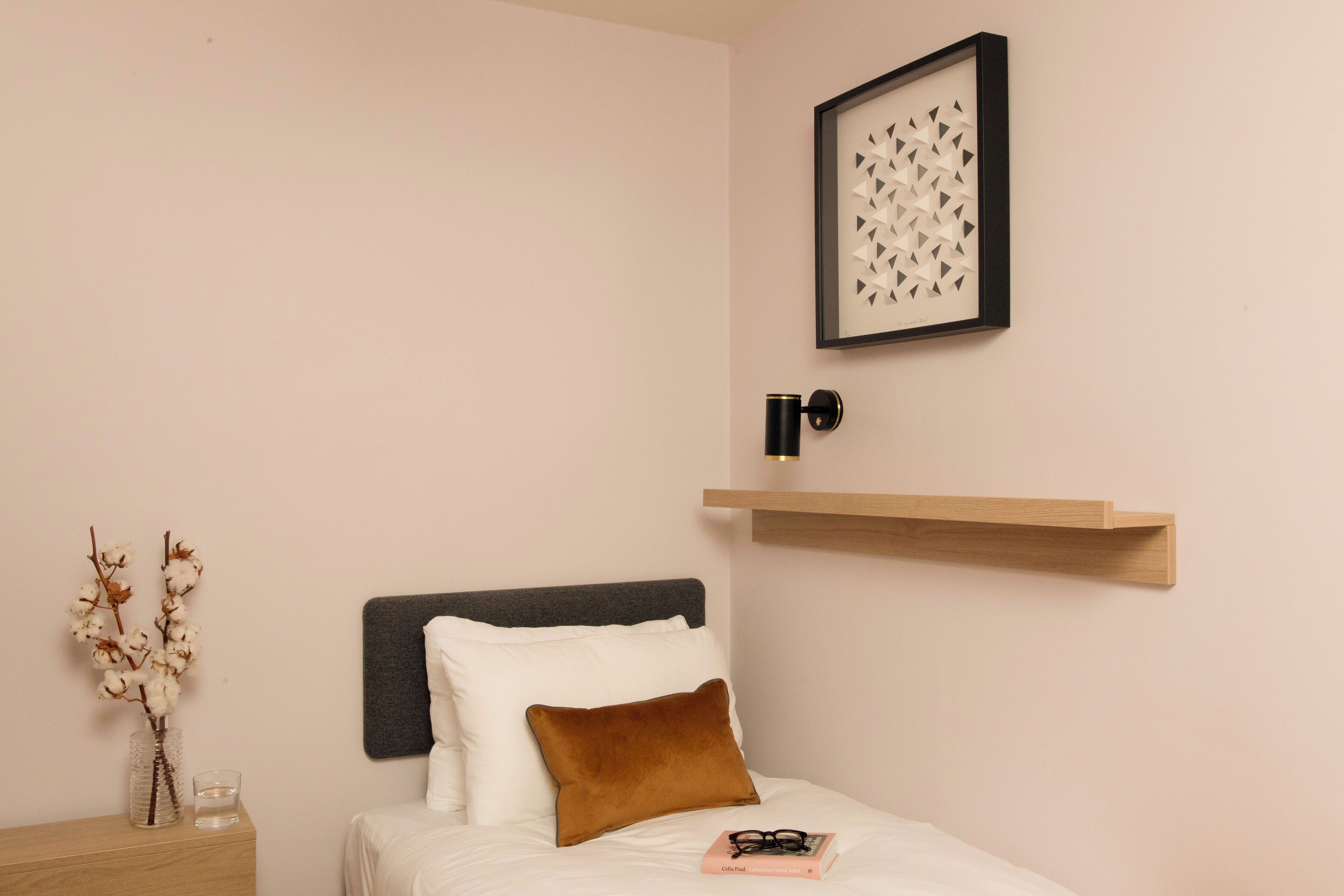Un dormitorio ordenado con cama, mesita de noche, un florero con flores secas, un cuadro enmarcado y un estante montado en la pared.