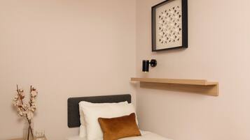 Een netjes ingerichte slaapkamer met een bed, een nachtkastje, een vaas met gedroogde bloemen, een ingelijst beeld en een wandplank.