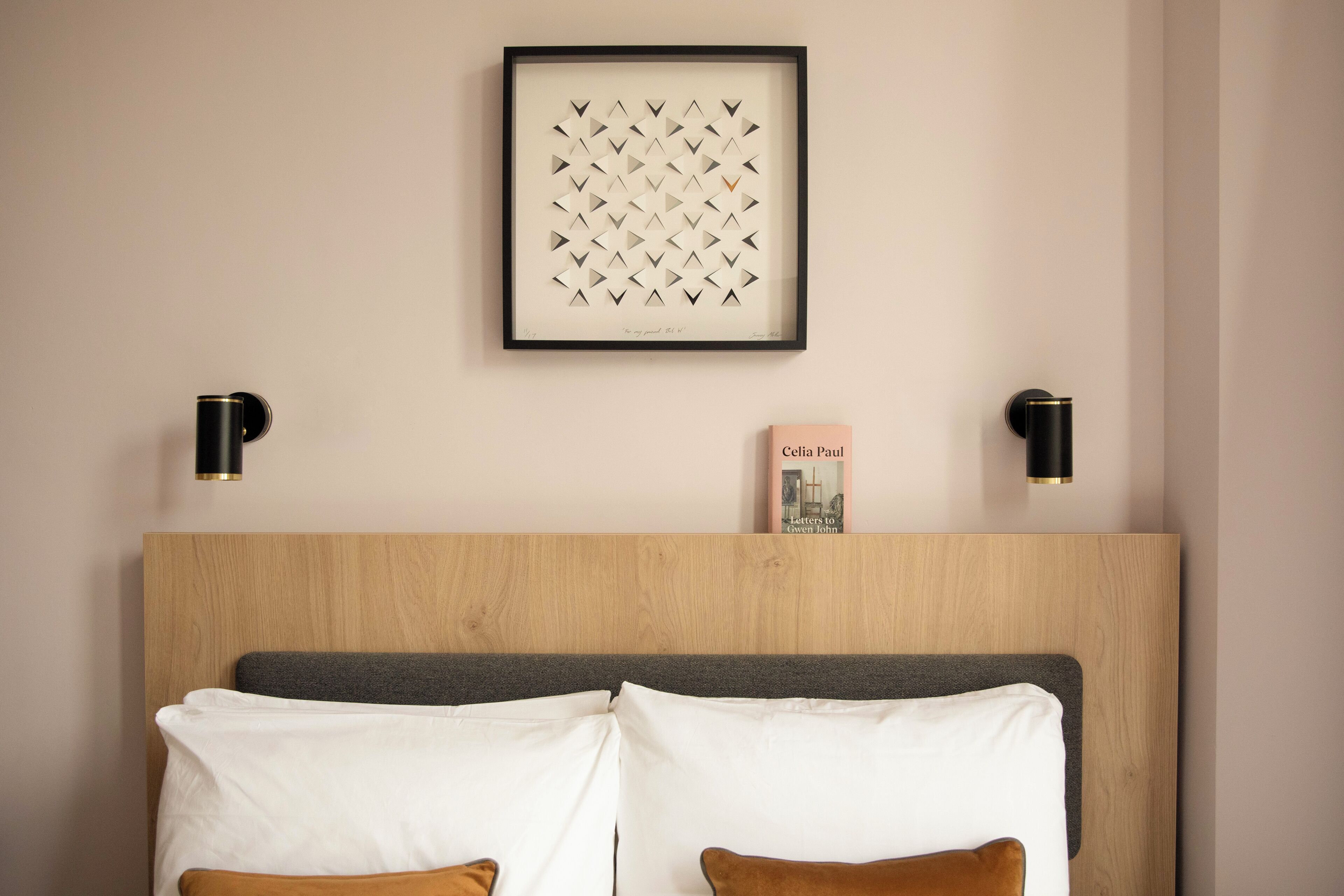 Une chambre d’hôtel bien aménagée, avec une tête de lit en bois, une œuvre d’art encadrée au-dessus du lit, et deux tables de chevet dotées de lampes noires.