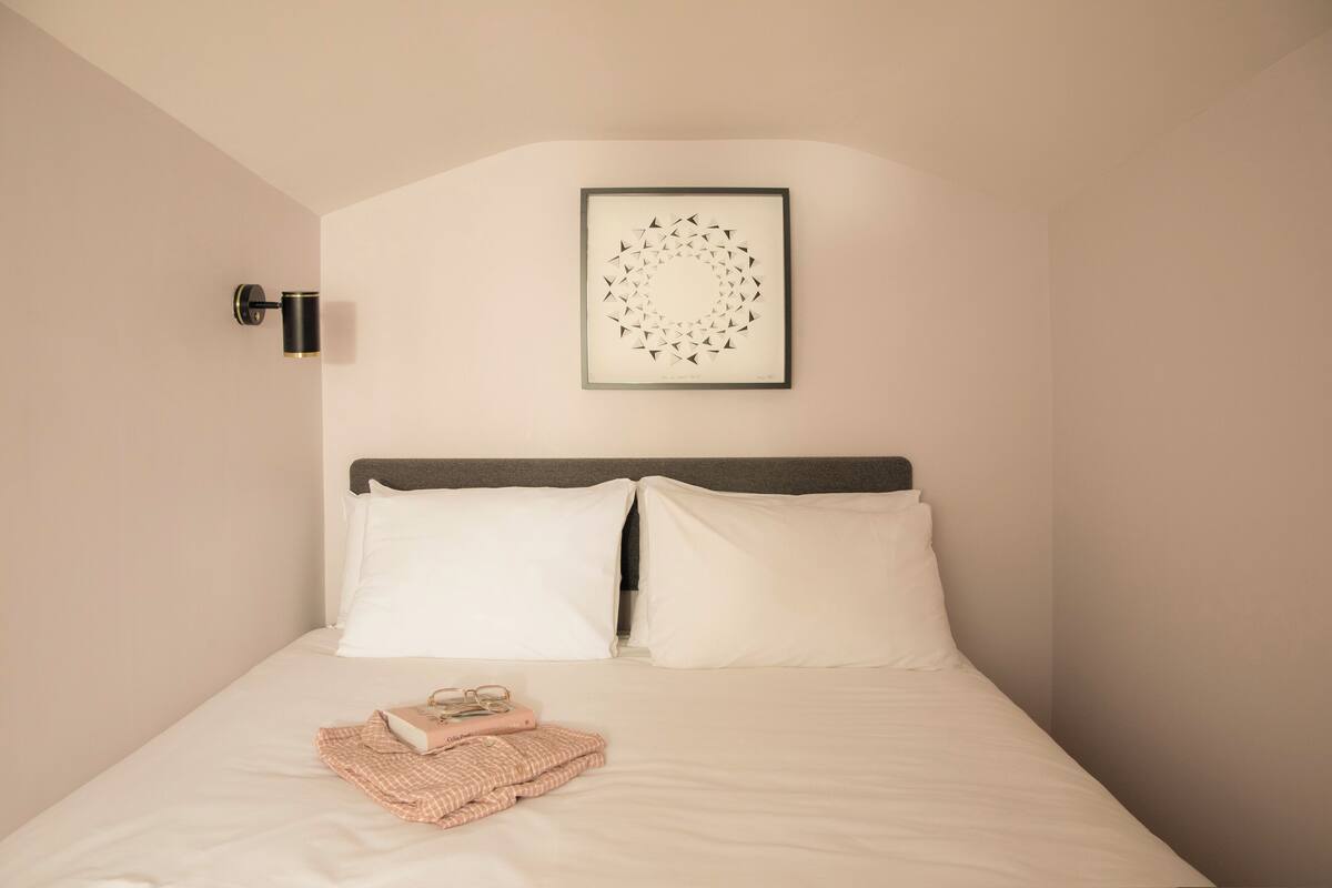 Een opgemaakt bed met witte lakens, een ingelijste afbeelding erboven en een nachtkastje met een lamp.