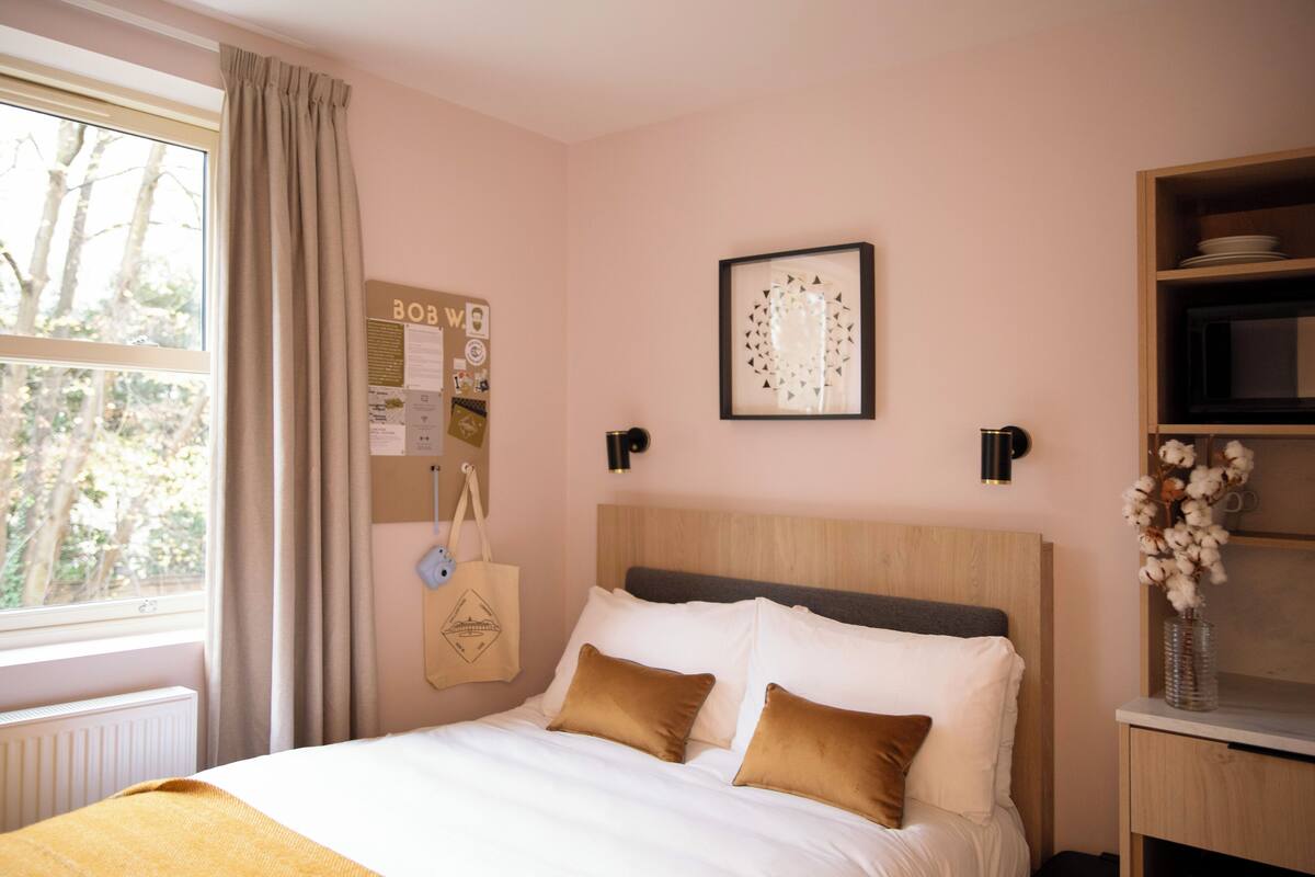 Een slaapkamer met een houten hoofdbord, een bed met wit en bruin beddengoed, een nachtkastje met een bloevas en een raam met gordijnen.