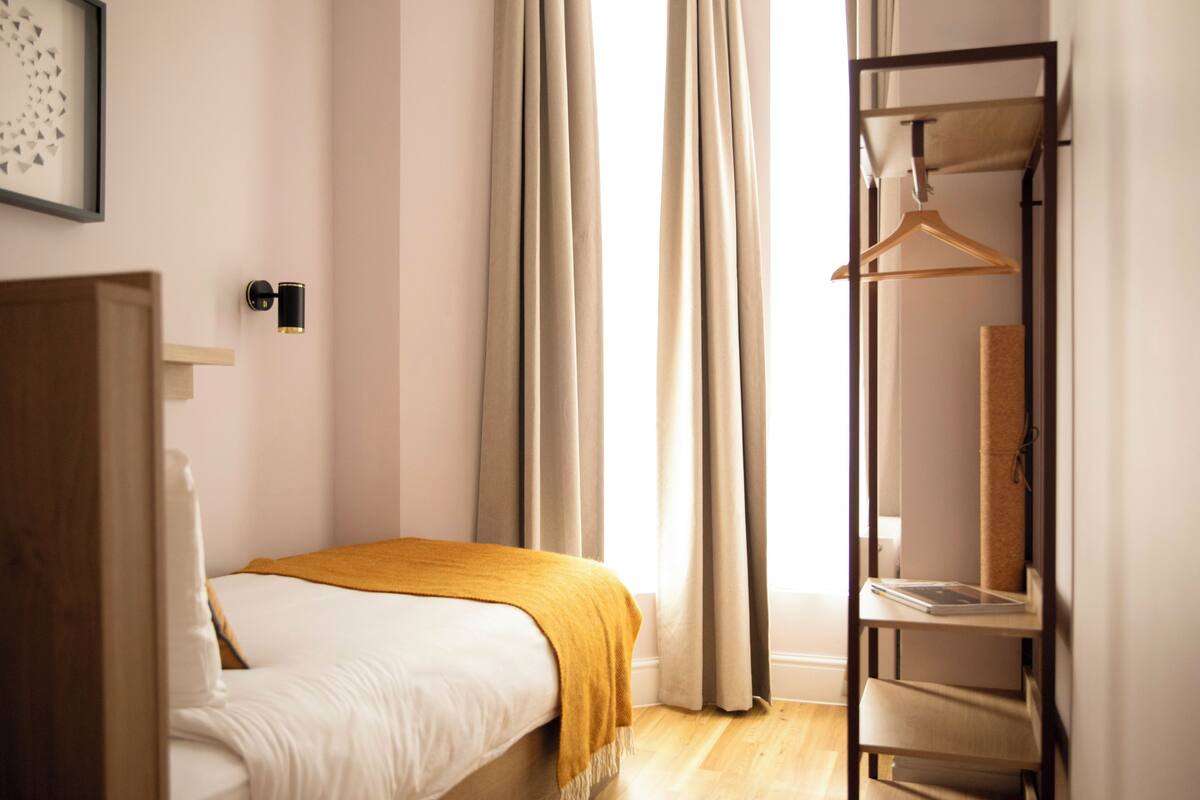 Een slaapkamer met een bed, een houten garderobe, een zwevende kast, een raam met gordijnen en een wandlamp.