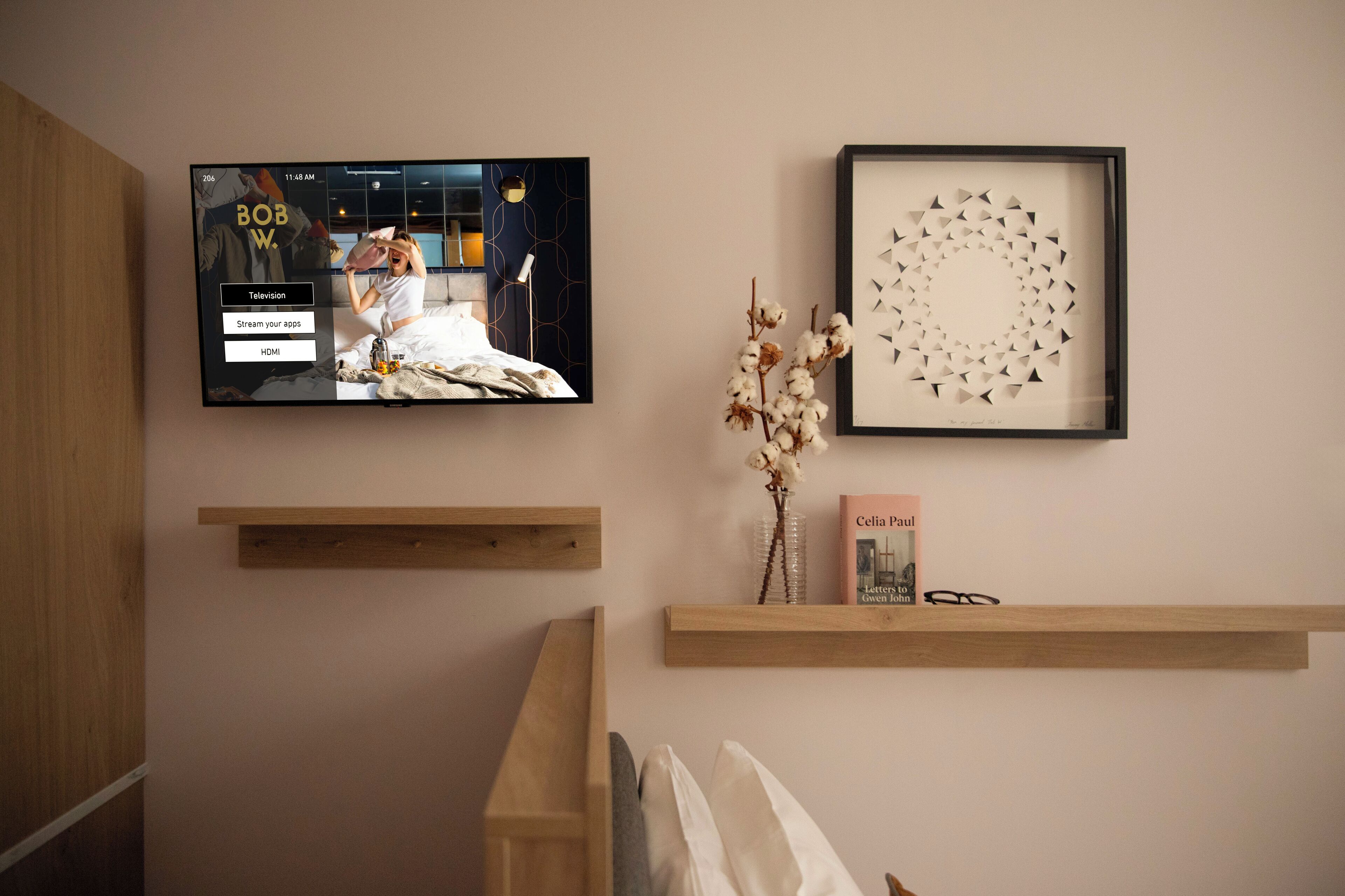 A TV mounted on a wall displaying a TV show, a framed abstract art piece, and a small shelf with a book and a vase.