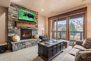 A living room with a stone fireplace, a television mounted on the wall, a large window with a view of mountains, and a cozy seating area.