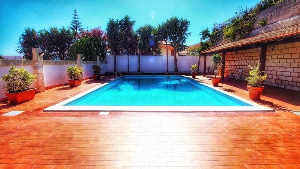 A rectangular swimming pool with a tiled deck, surrounded by potted plants and a white wall. - Vacation home with dog boarding (Messina)