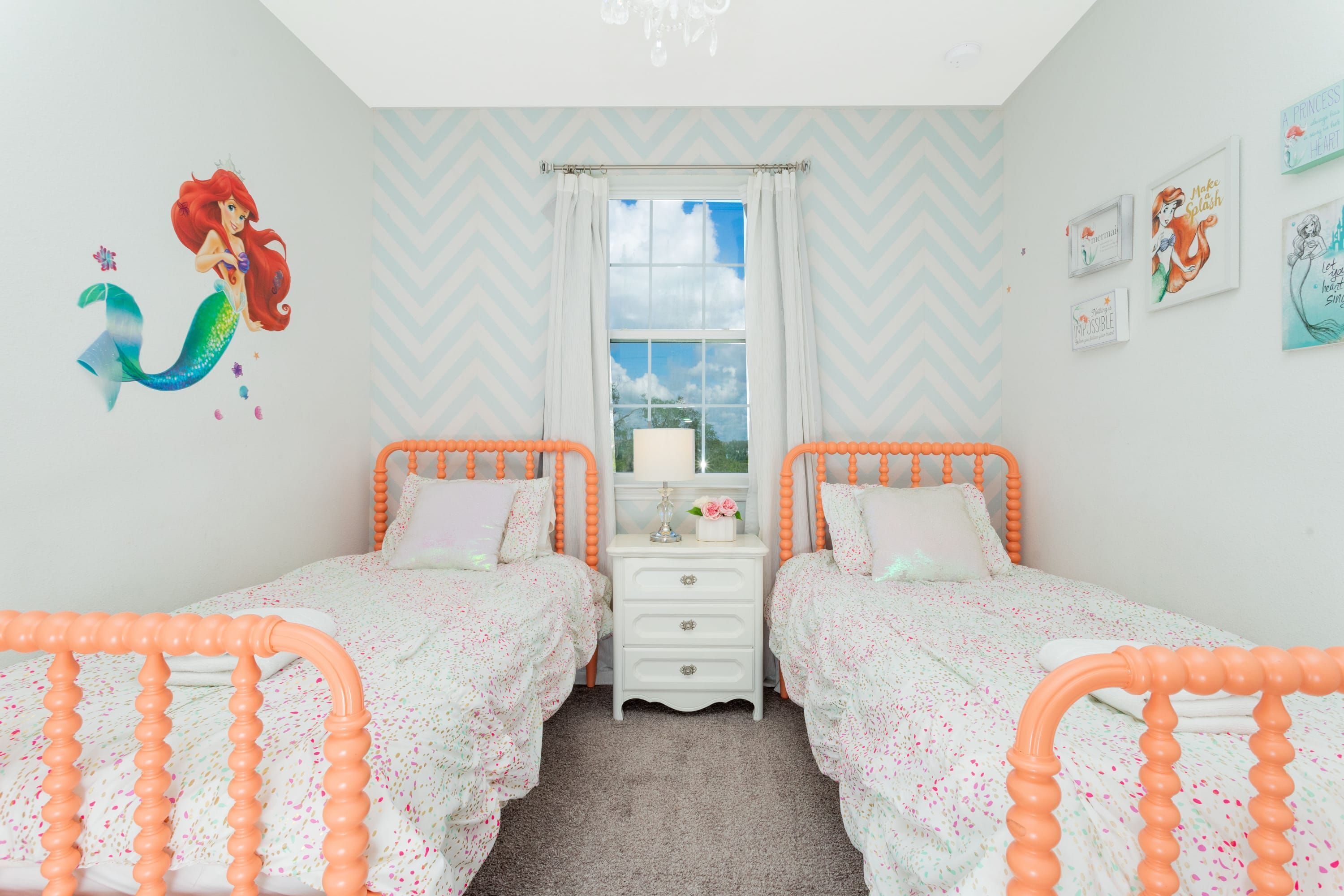 A bedroom with two beds, a dresser, a window with curtains, and wall decorations including a mermaid mural and framed pictures.