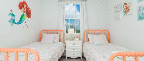 A bedroom with two beds, a dresser, a window with curtains, and wall decorations including a mermaid mural and framed pictures.