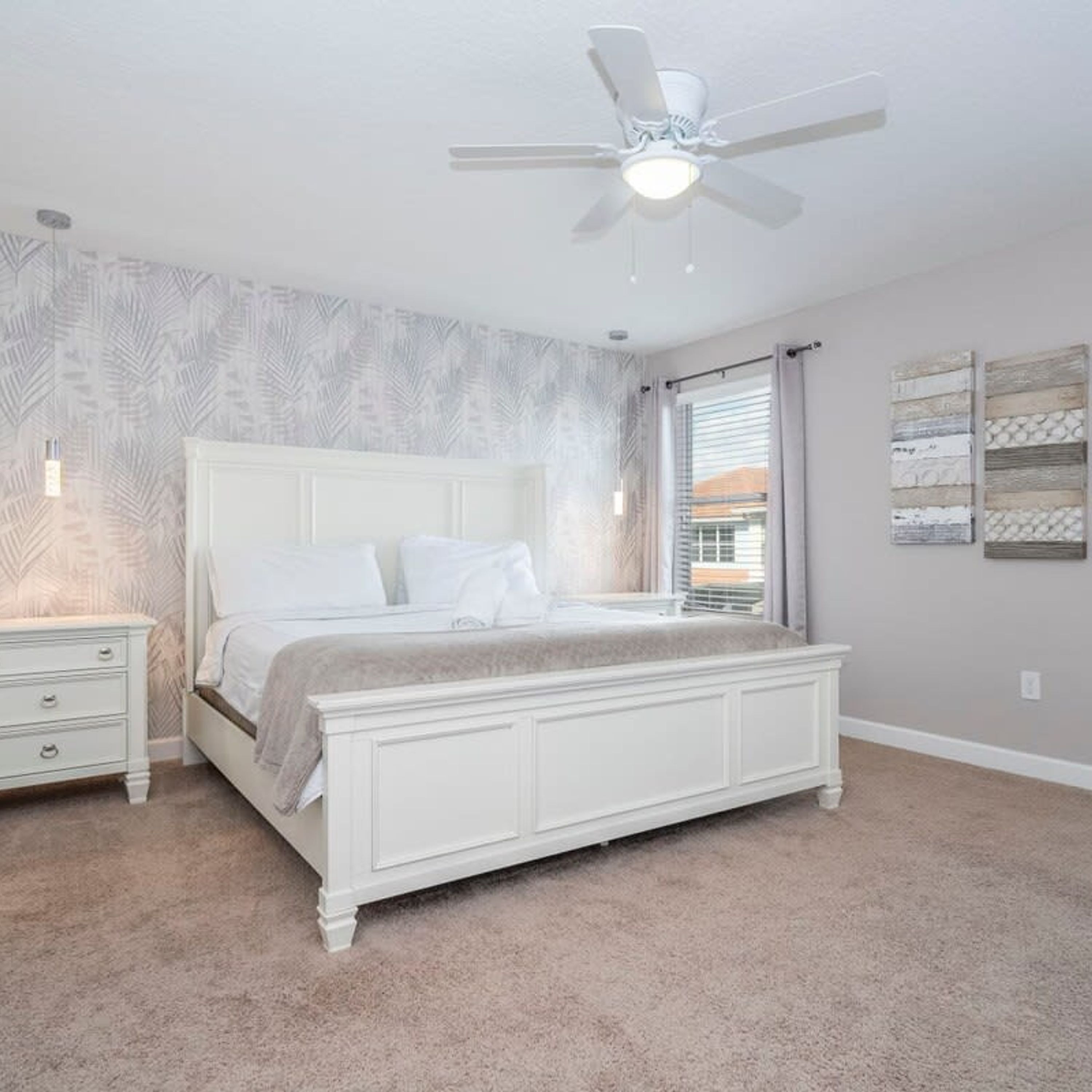Une chambre à coucher avec un lit blanc, une table de chevet, un ventilateur de plafond et une fenêtre avec des rideaux.