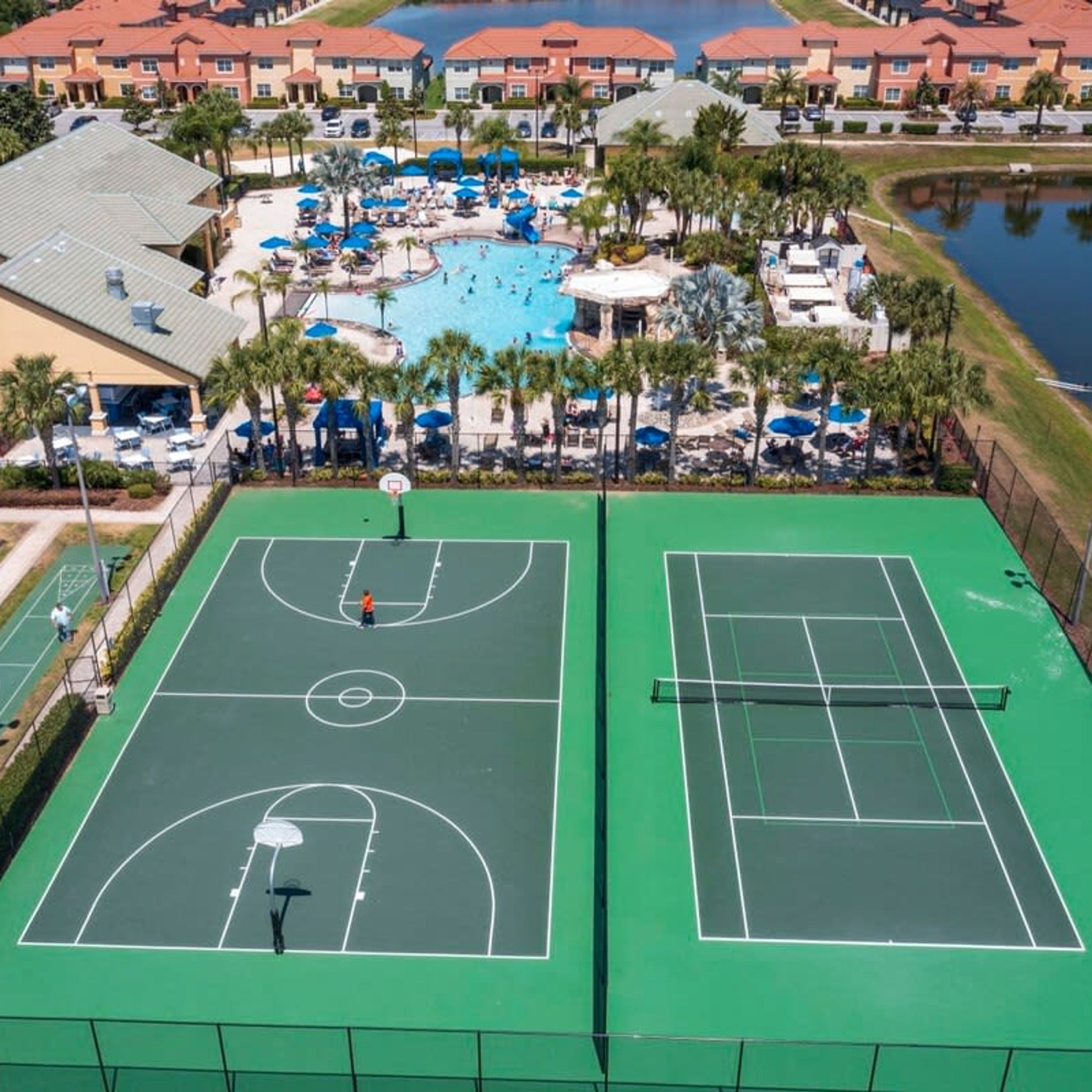 A tennis court with a basketball court adjacent to it, surrounded by a pool area with umbrellas and palm trees.