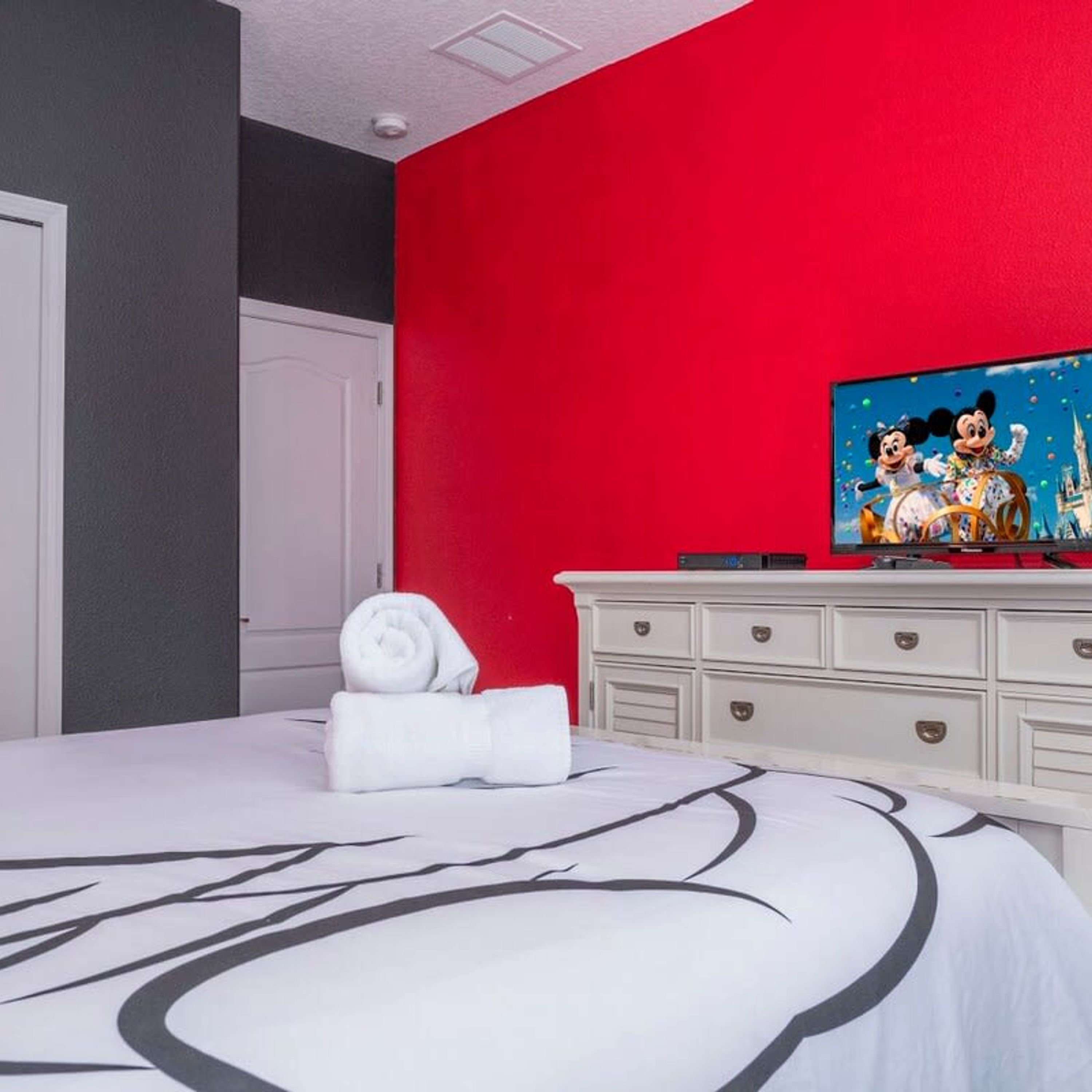 Een hotelkamer met een rode accentmuur, een bed met een zwart-wit ontwerp, een witte dressoir en een televisie waarop geanimeerde personages te zien zijn.