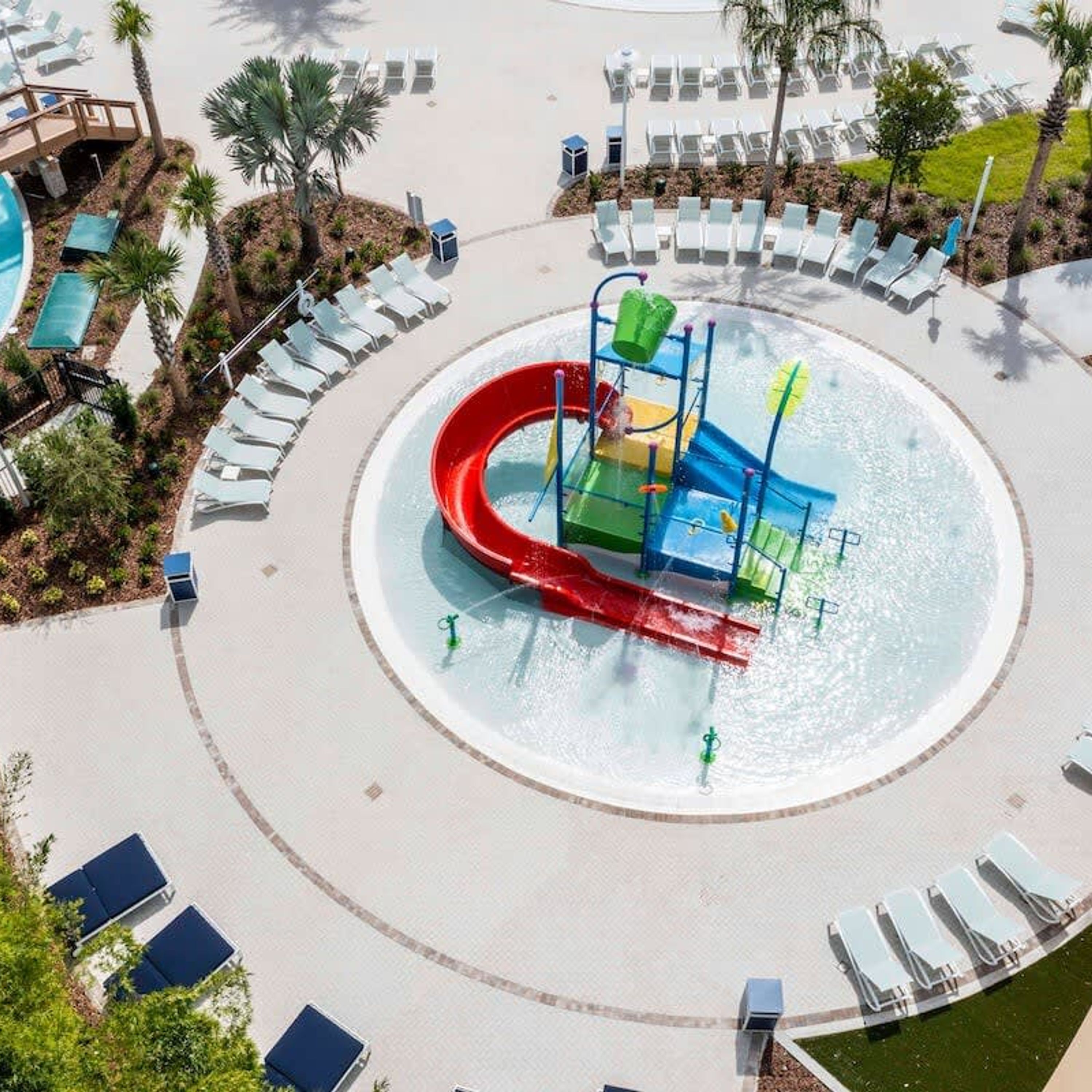 A water park with a central water slide, surrounded by lounge chairs and palm trees.