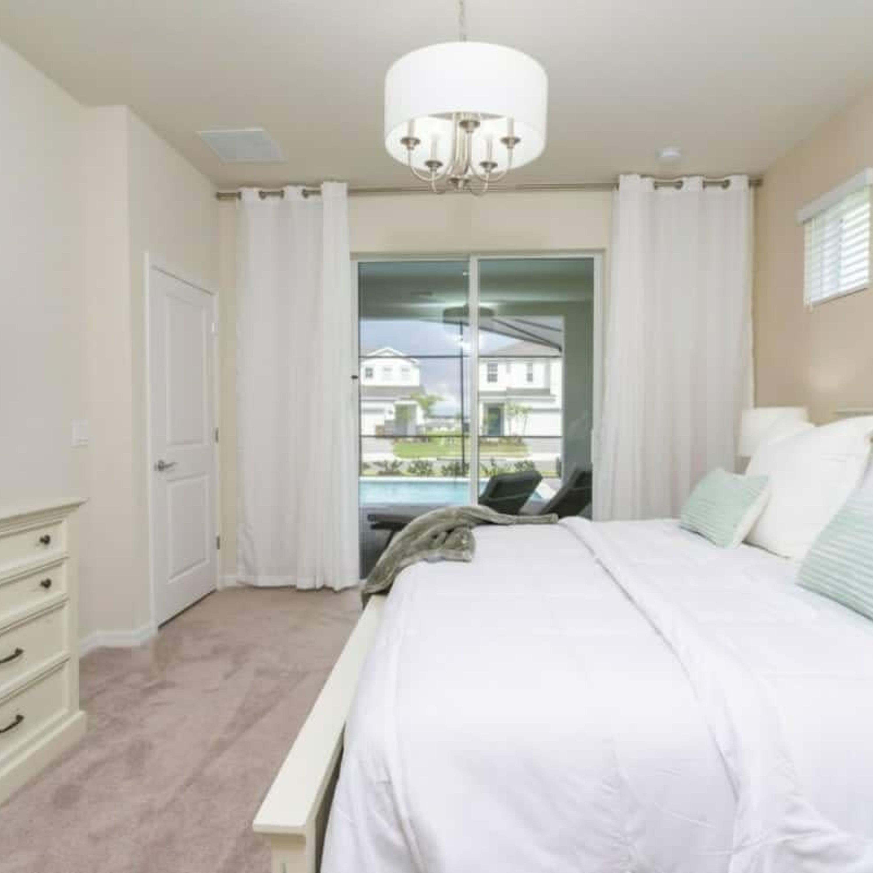 A bedroom with a large bed, a bench, a chandelier, and a view of a house through a glass door.