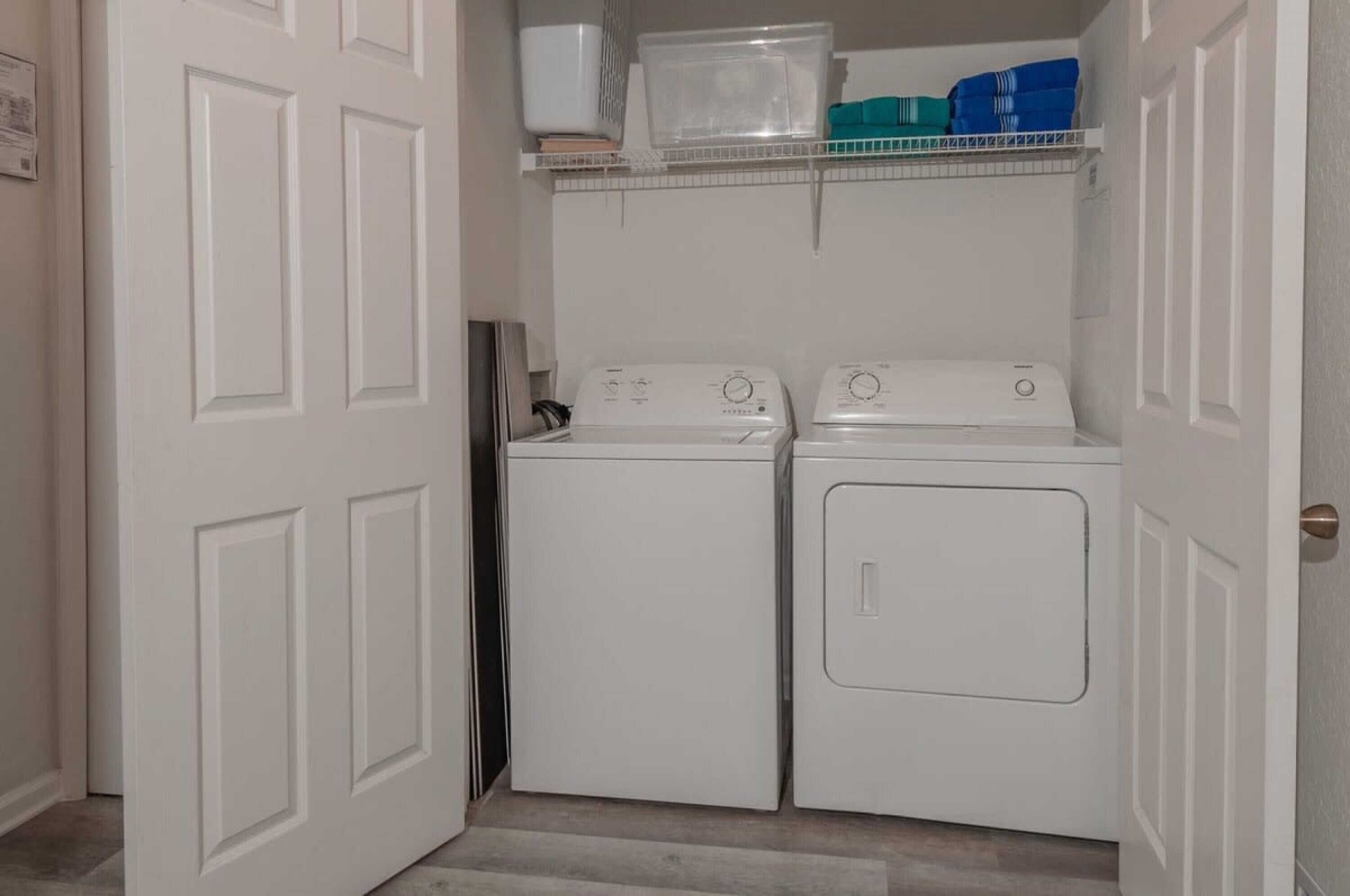 A laundry room with two white front-loading washers and dryers, a white door, and a shelf above holding a plastic container and folded towels.