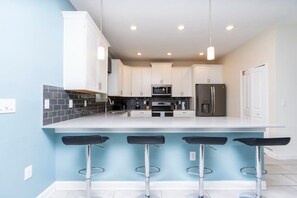A modern kitchen with white cabinets, a central island with bar stools, a microwave, and a refrigerator. - 8912 Amazing 6BD Home (Davenport)