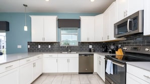 A modern kitchen with white cabinets, a black tiled backsplash, stainless steel appliances, and a window with blinds. - 8912 Amazing 6BD Home (Davenport)