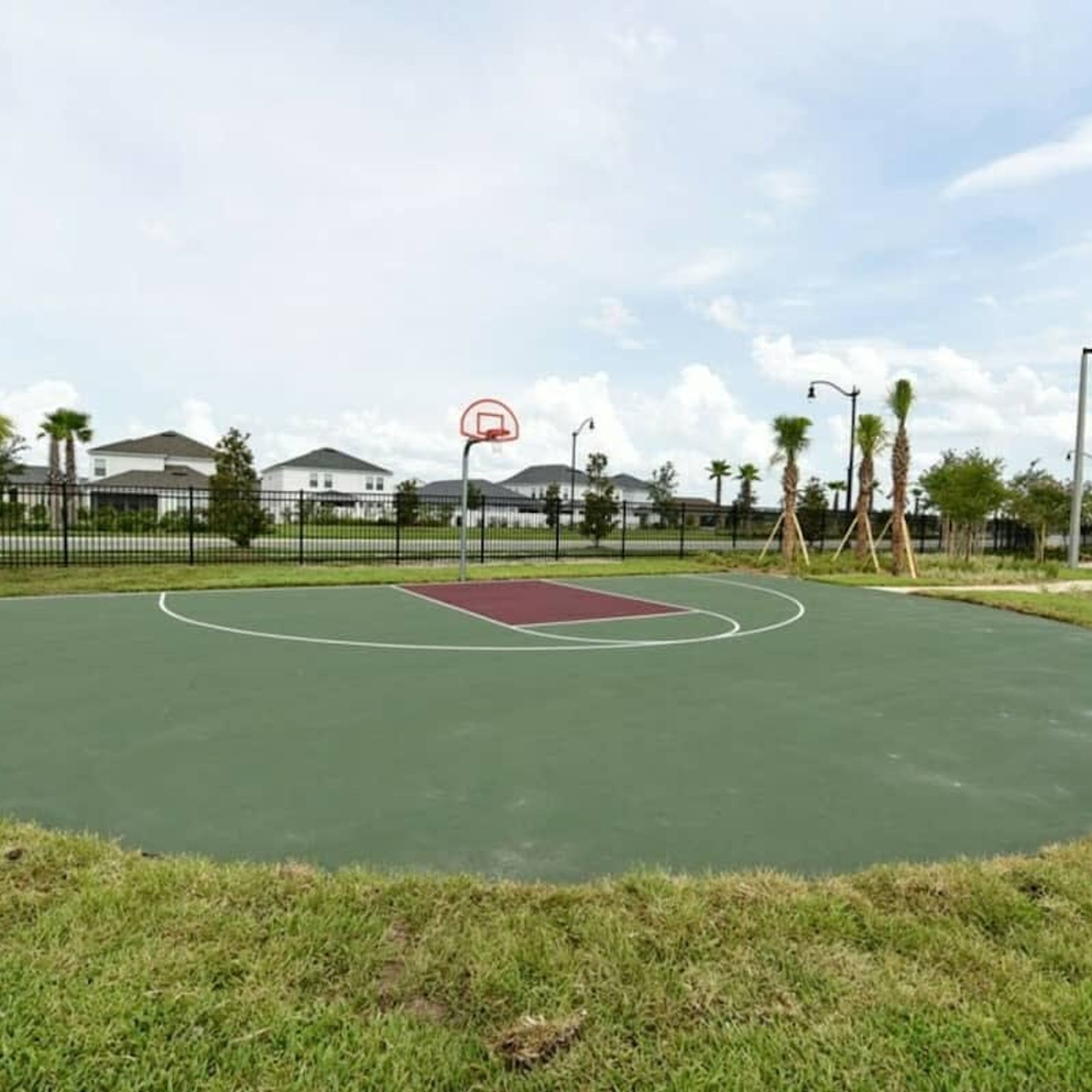 Een buitenbasketbalveld met een rode speeloppervlakte, omringd door een zwarte hek, palmbomen en huizen op de achtergrond.