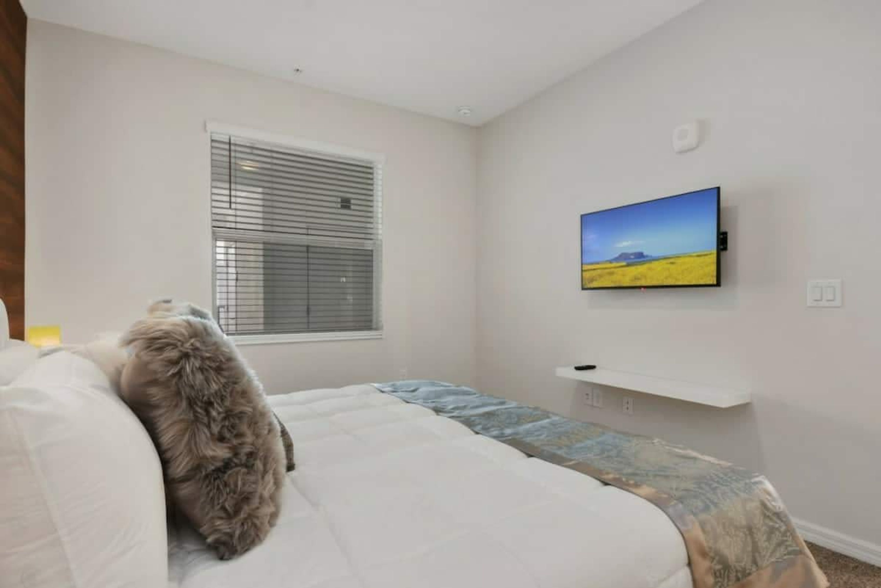 Un dormitorio con cama, una ventana con persianas, un televisor montado y una pequeña estantería.