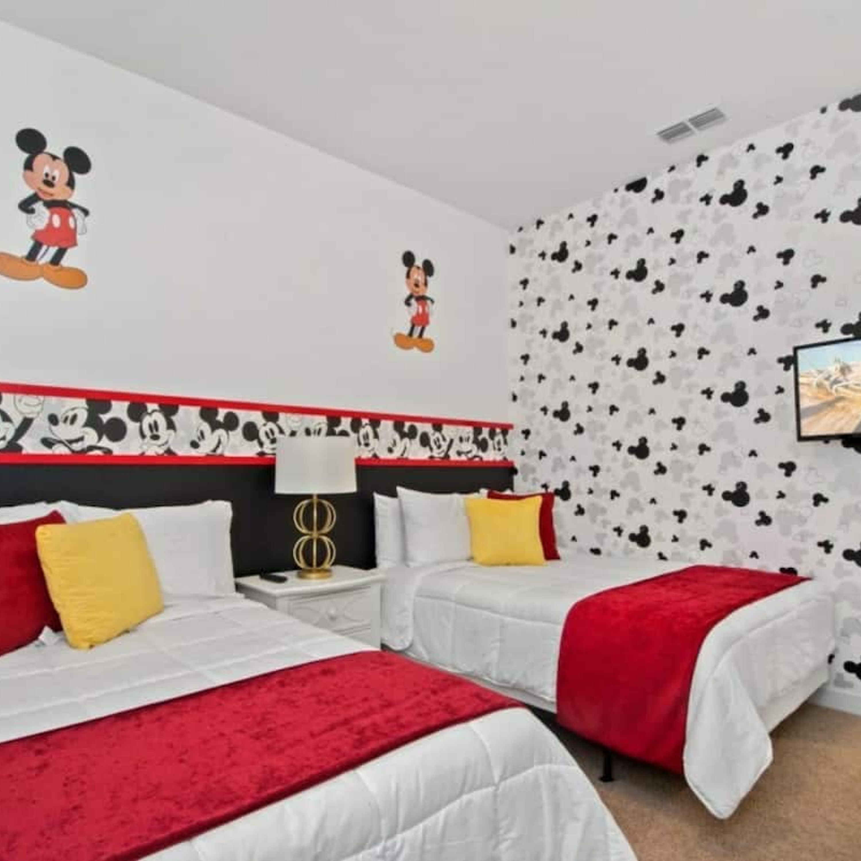 Habitación de hotel con dos camas, cada una con ropa de cama roja y blanca, un adhesivo de pared de Mickey Mouse y un papel tapiz con estampado blanco y negro.
