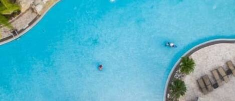 Vista aérea de una piscina con un diseño ondulado distintivo, rodeada de zonas de descanso con sillas y sombrillas, y un pequeño elemento acuático.