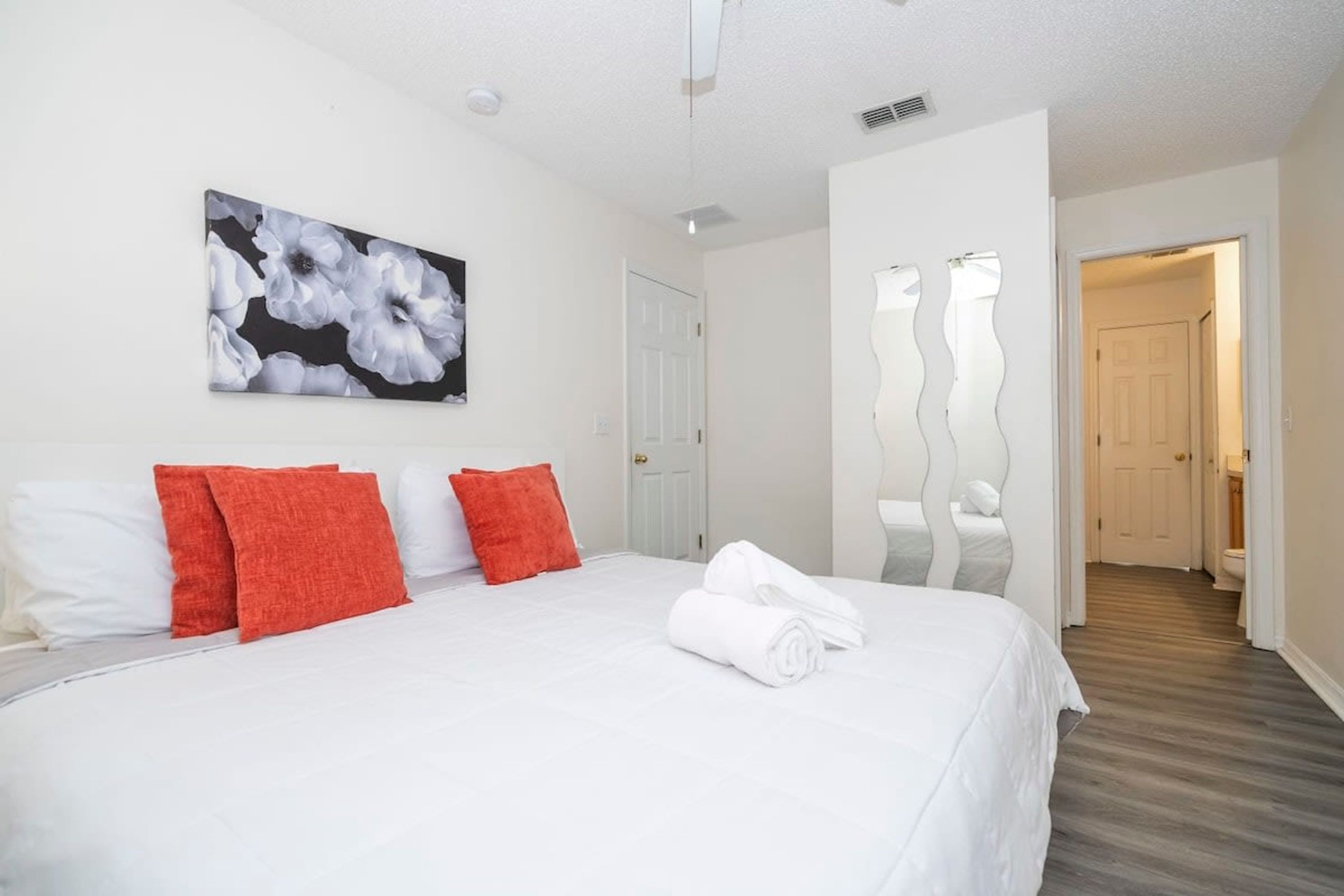 Ein Hotelzimmer mit einem großen Bett, weißer Bettwäsche, roten Kissen, einem Blumenbild an der Wand und einem Gang zu einem weiteren Zimmer.
