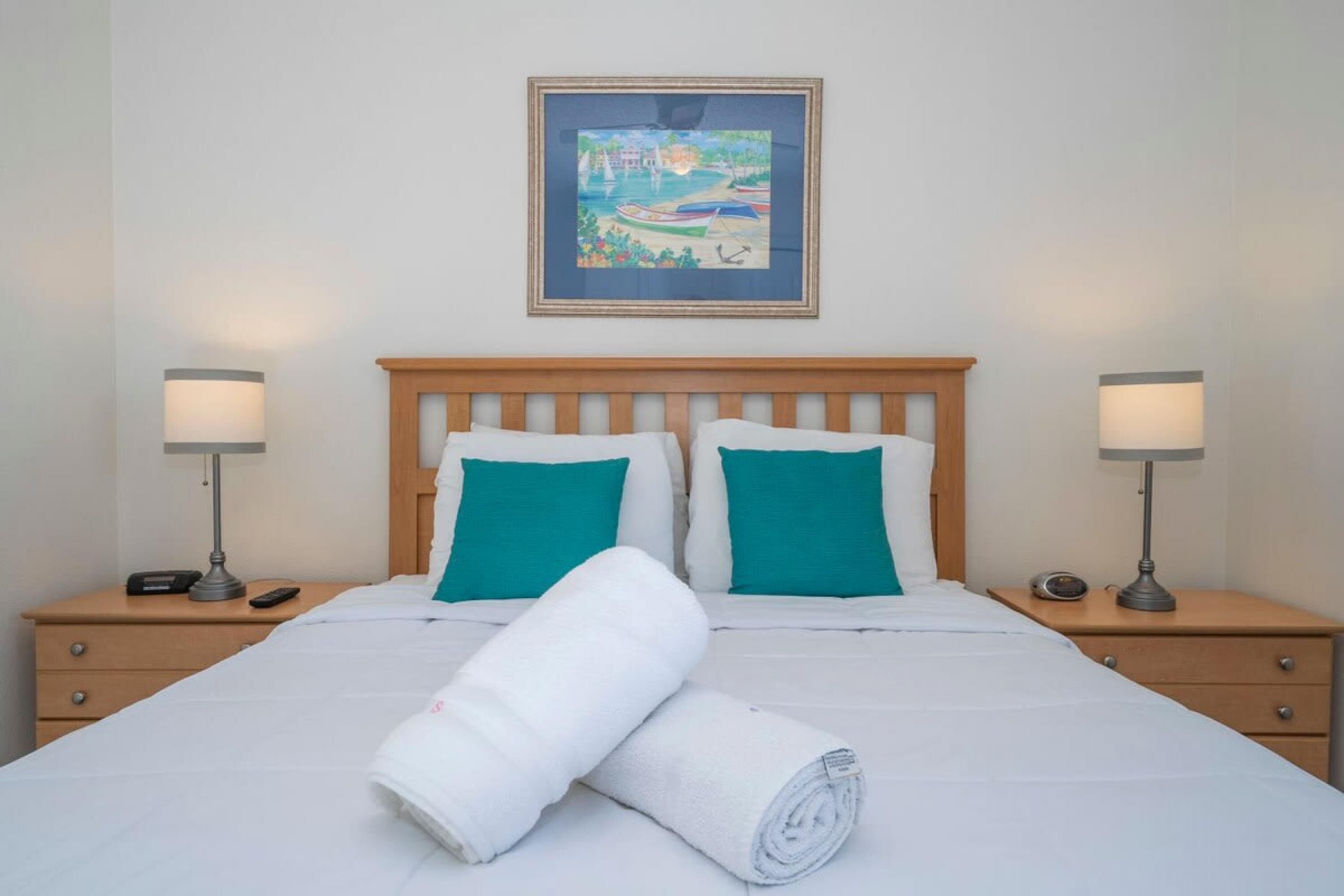 Een netjes opgemaakt bed met wit beddengoed en blauwgroene kussens, met nachtkastjes met lampen ernaast en een ingelijst schilderij erboven.