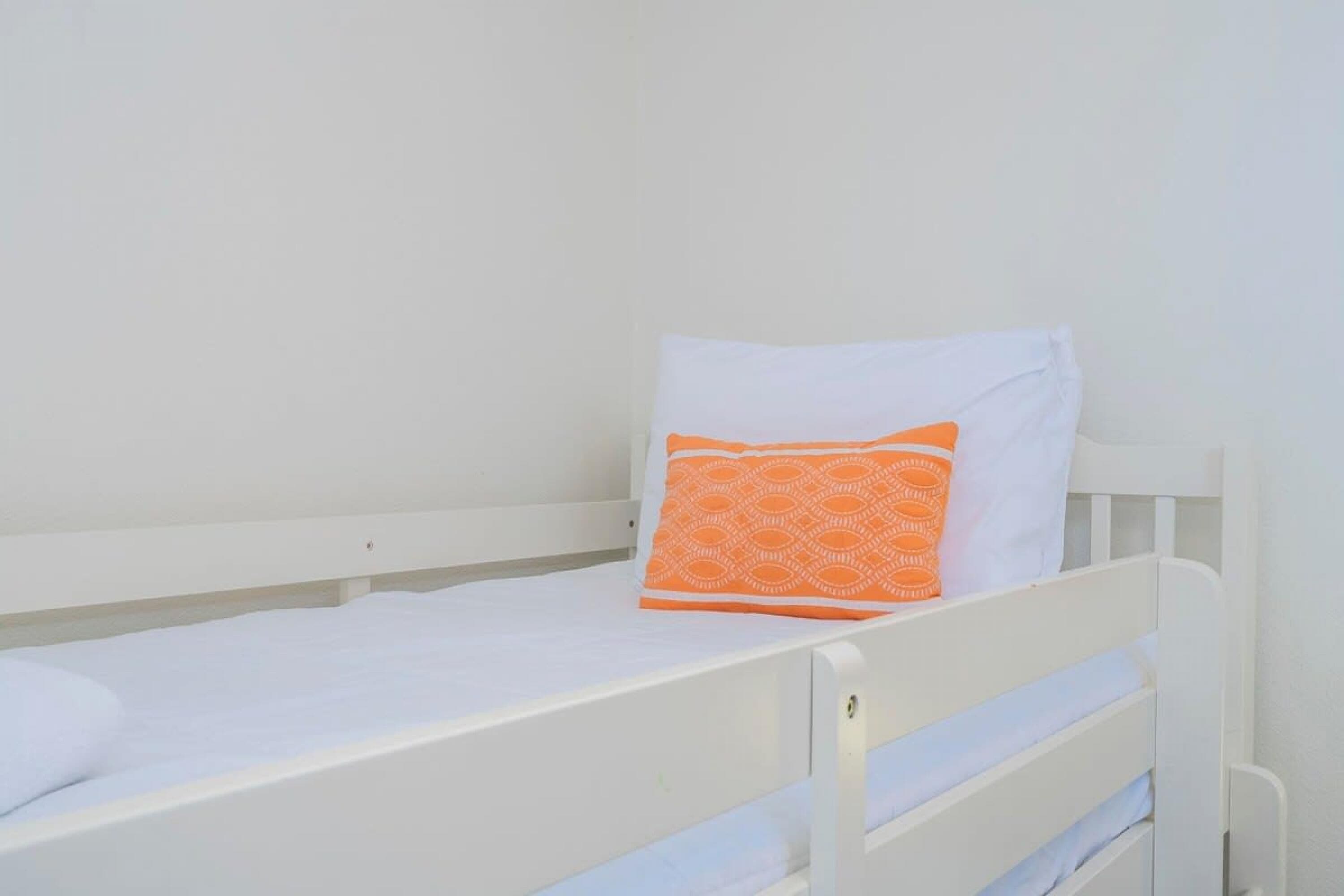 A single white bed with an orange patterned pillow and a white pillow.