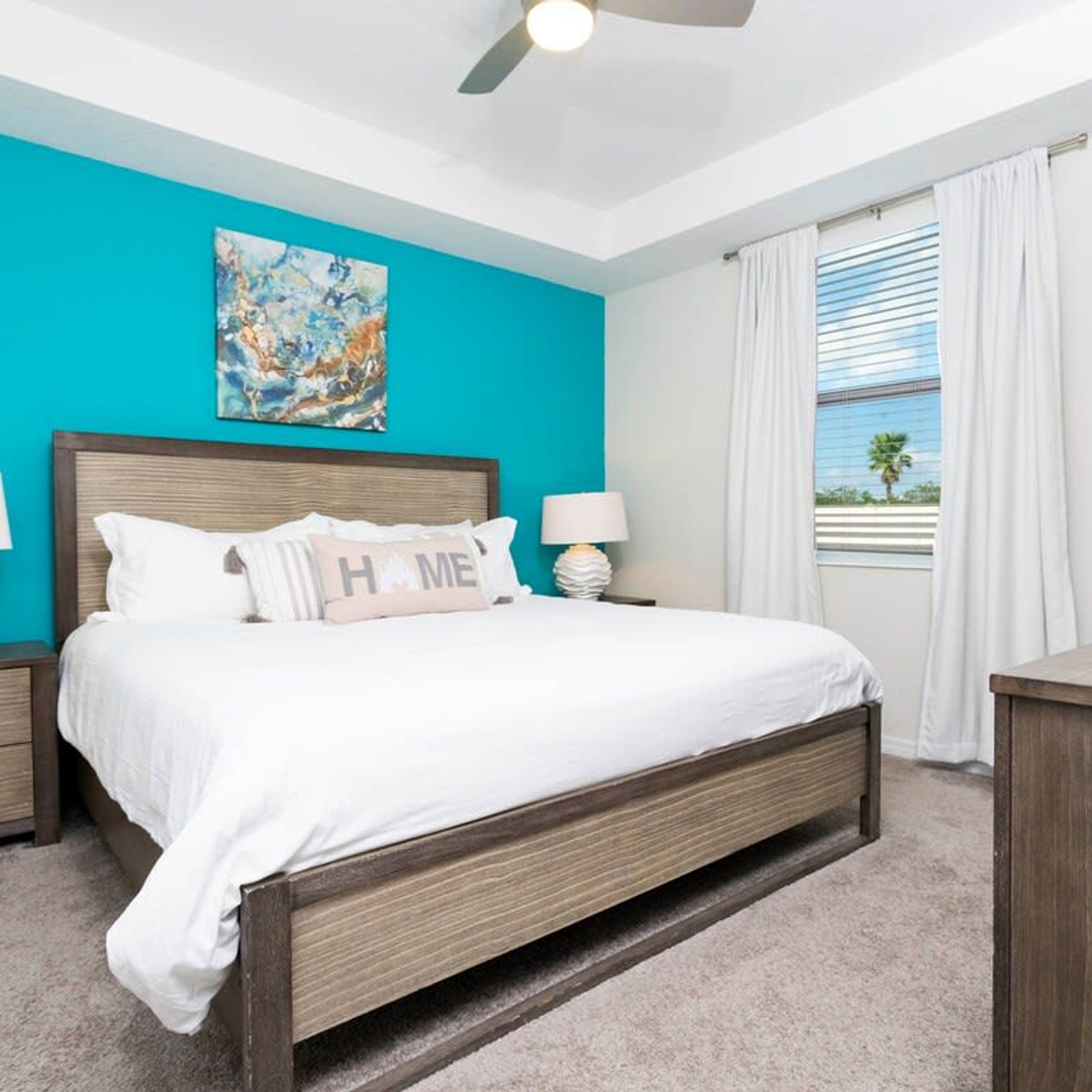 Une chambre à coucher avec un lit, un ventilateur de plafond, une table de chevet, une lampe, un tableau et une fenêtre avec des rideaux.
