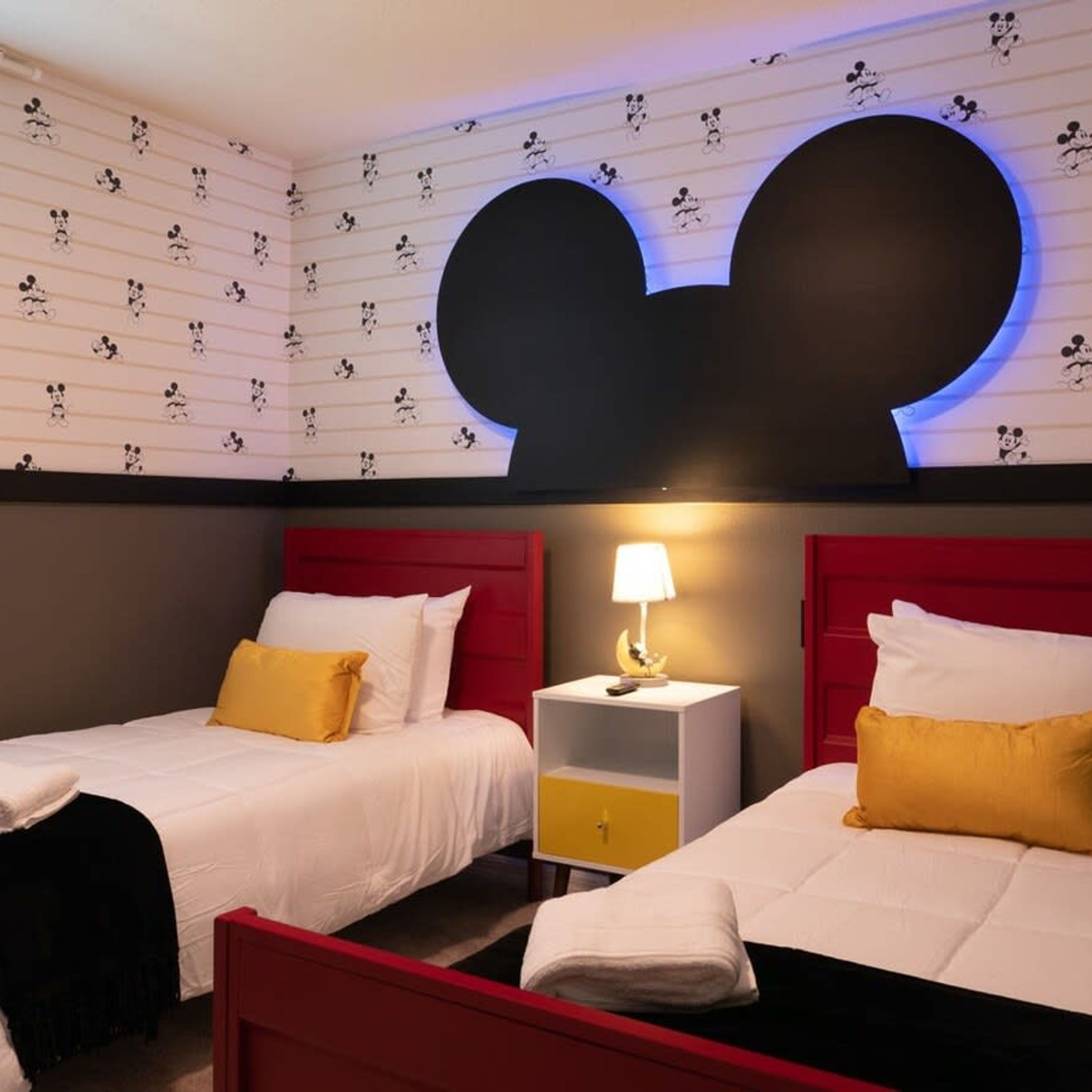 A hotel room with two beds, each with white bedding and yellow pillows. There is a large Mickey Mouse head decoration on the wall above the beds.
