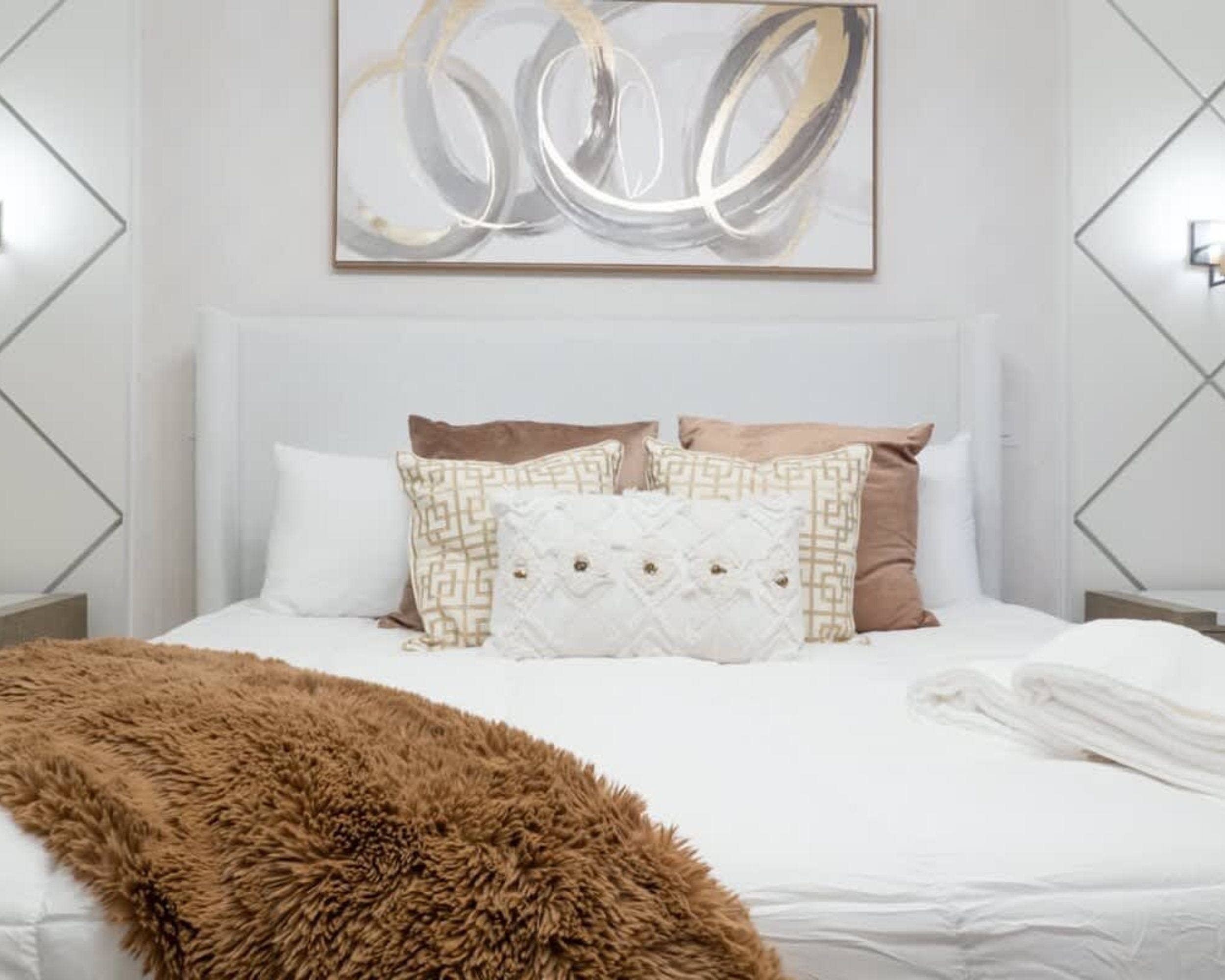 A modern bedroom with a large bed, decorative pillows, a fluffy throw blanket, and a geometric abstract painting on the wall.