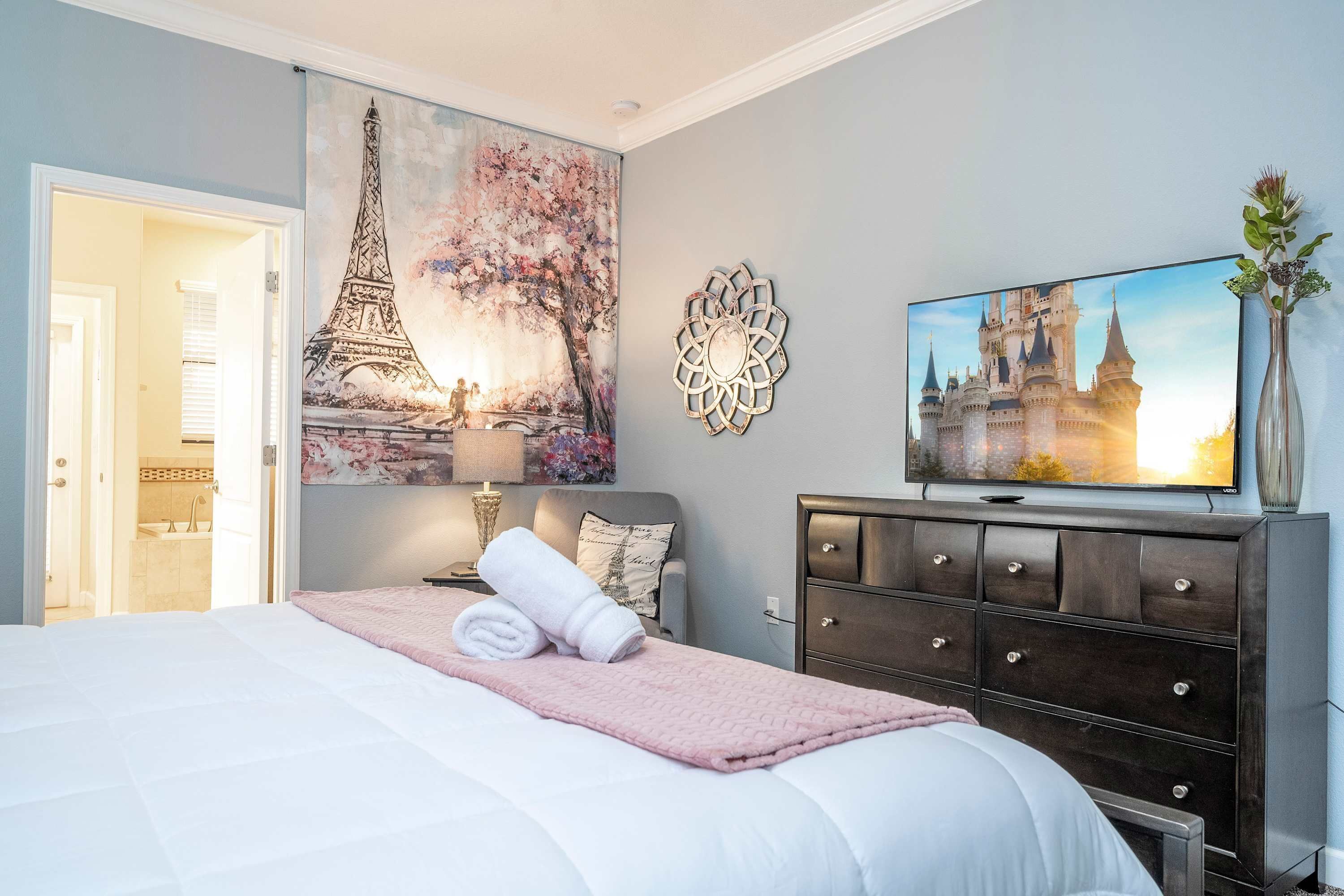 Een net opgemaakt bed met een roze plaid, een televisie op een dressoir en een schilderij van de Eiffeltoren.