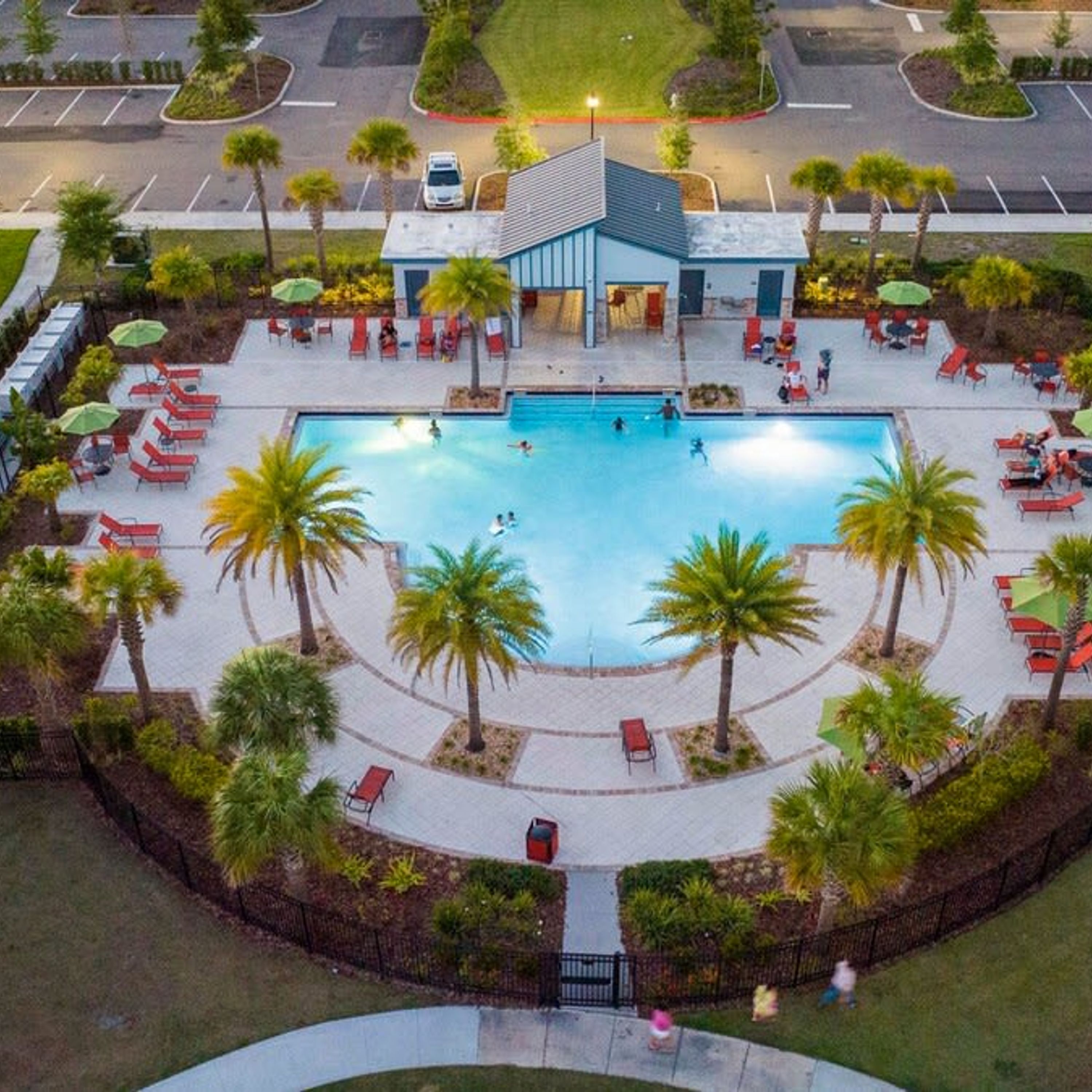 An aerial view of a resort with a central swimming pool, surrounded by palm trees and red lounge chairs.