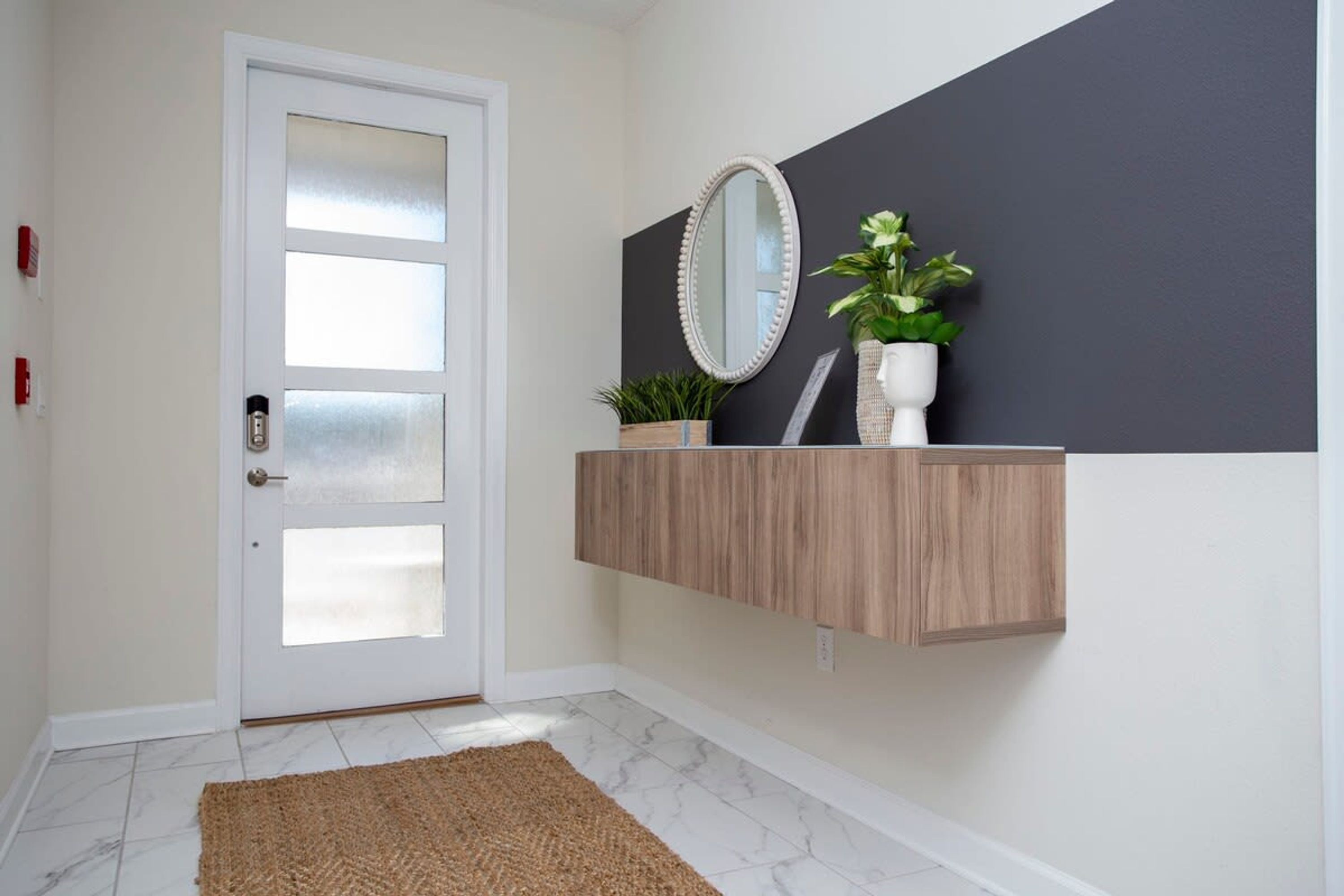 A hallway with a white door, a wooden floating shelf with a mirror and plants, and a marble floor.