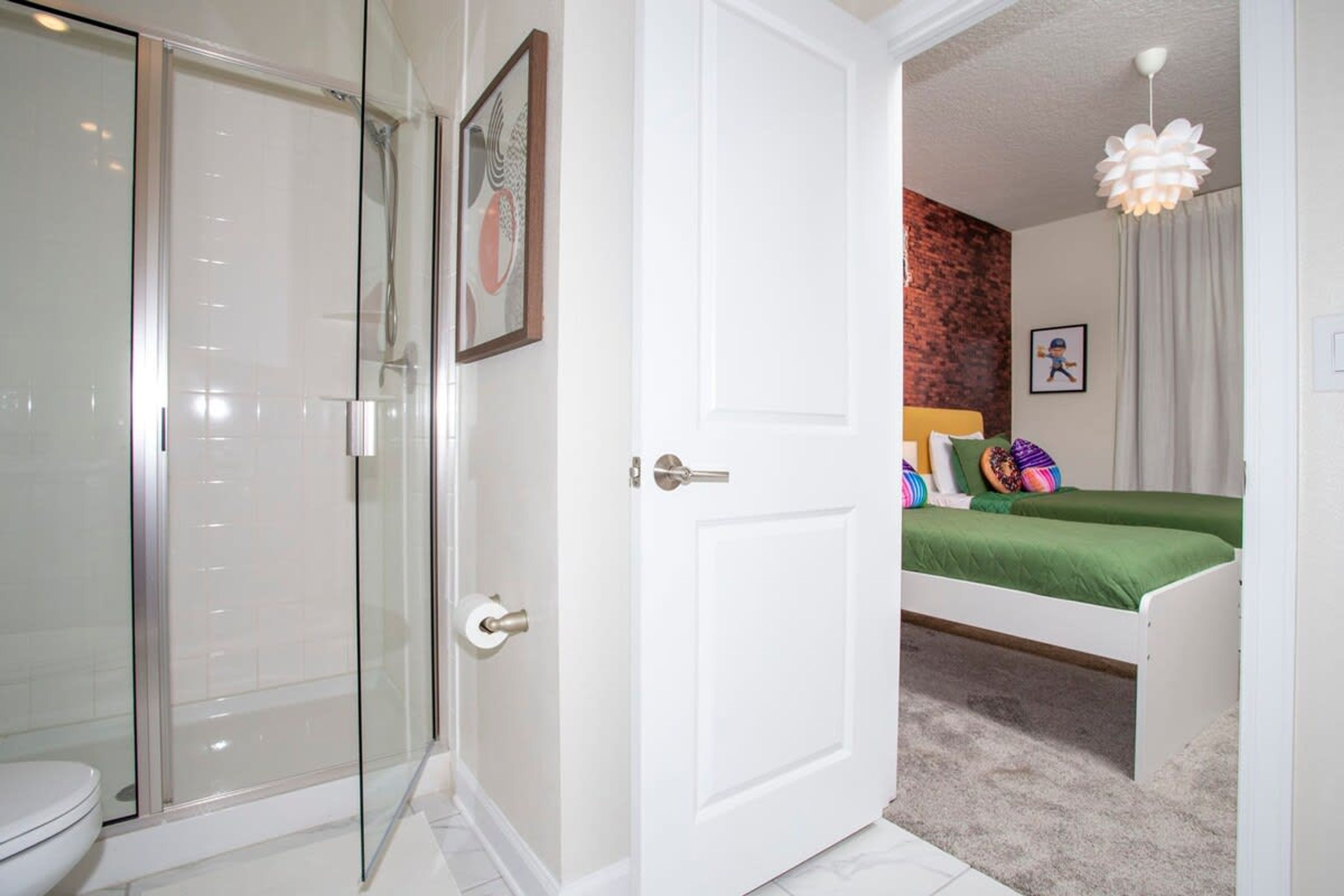 A bathroom with a glass shower enclosure, a toilet, and a white door leading to a bedroom with a bed and a headboard.