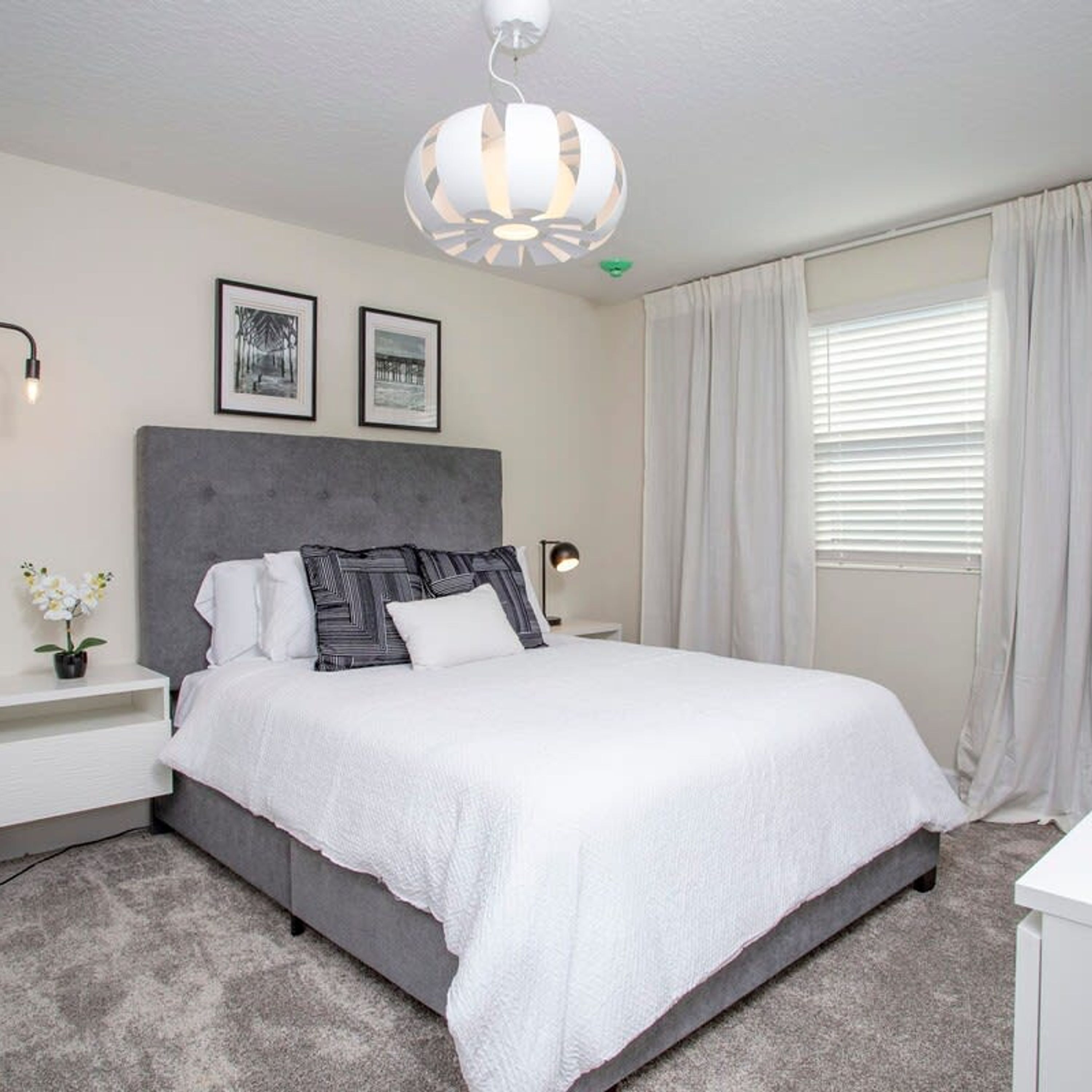 A bedroom with a large bed, a grey headboard, white bedding, a bedside table with a lamp, and a window with curtains.