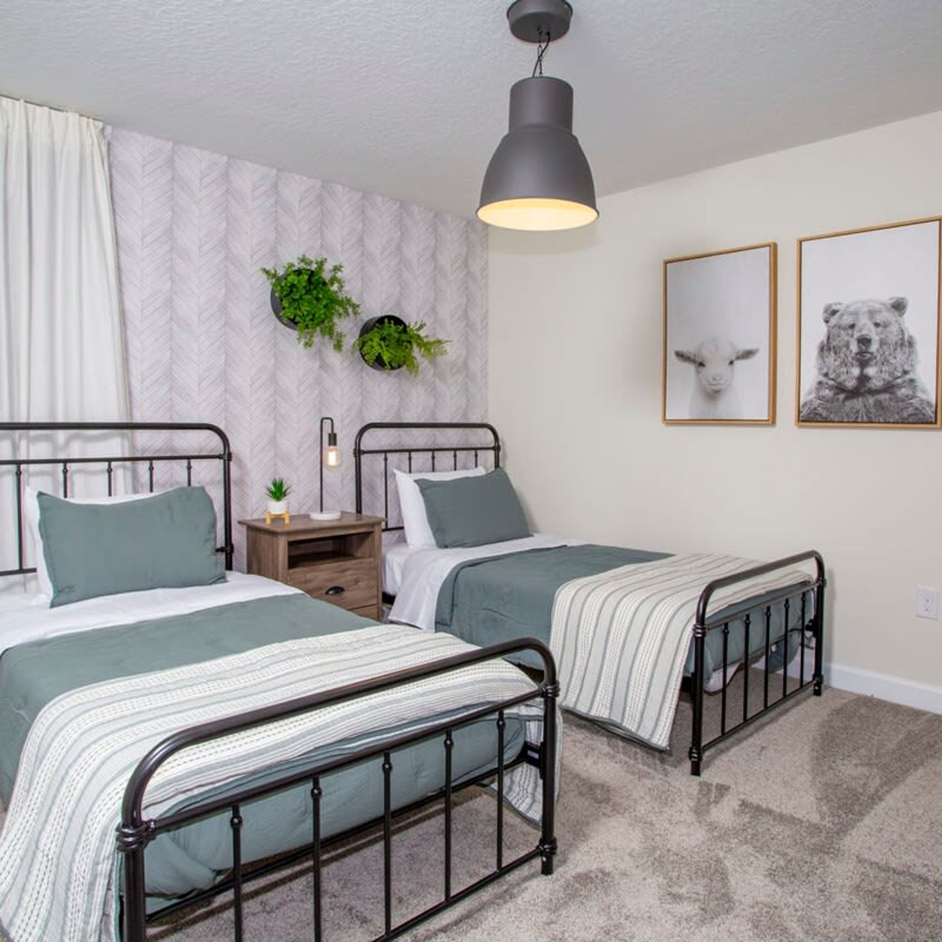 A bedroom with two metal beds, a nightstand with a lamp, a wall-mounted plant, and two framed pictures.