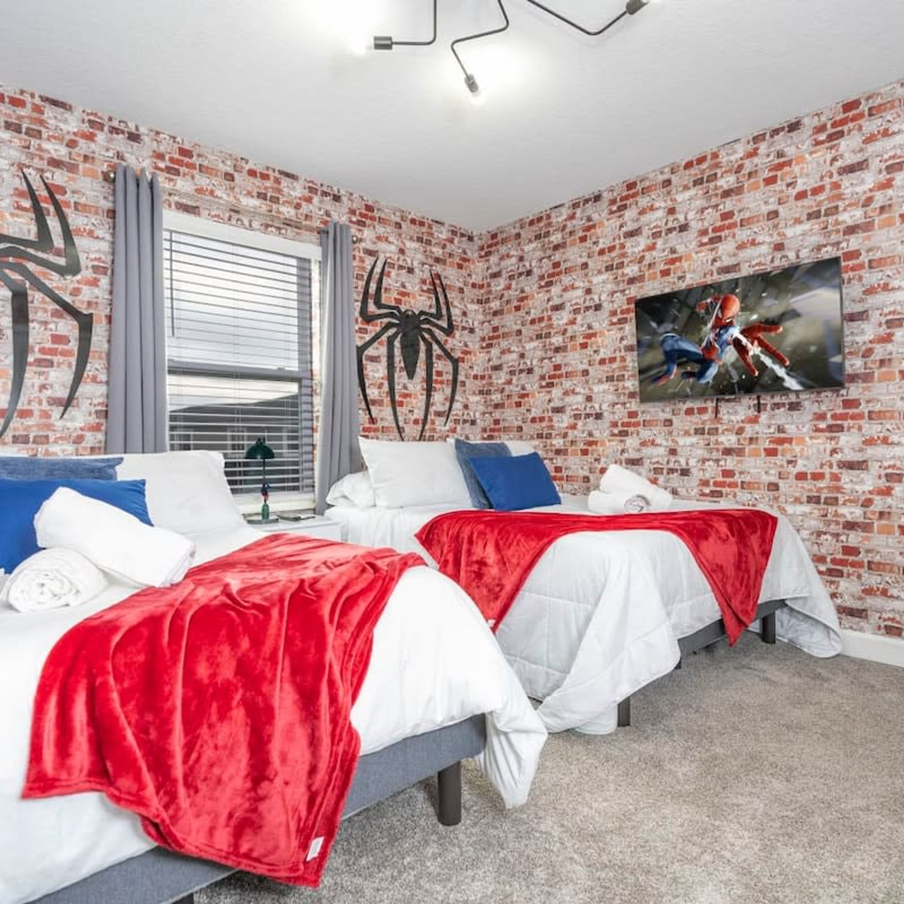 Ein Hotelzimmer mit einer Wand im Ziegelsteinmuster, zwei Betten mit rot-weißer Bettwäsche, einer großen Spinngespinst-Kunst und einem Spider-Man-Fernseher, sowie einem Fenster mit Jalousien.