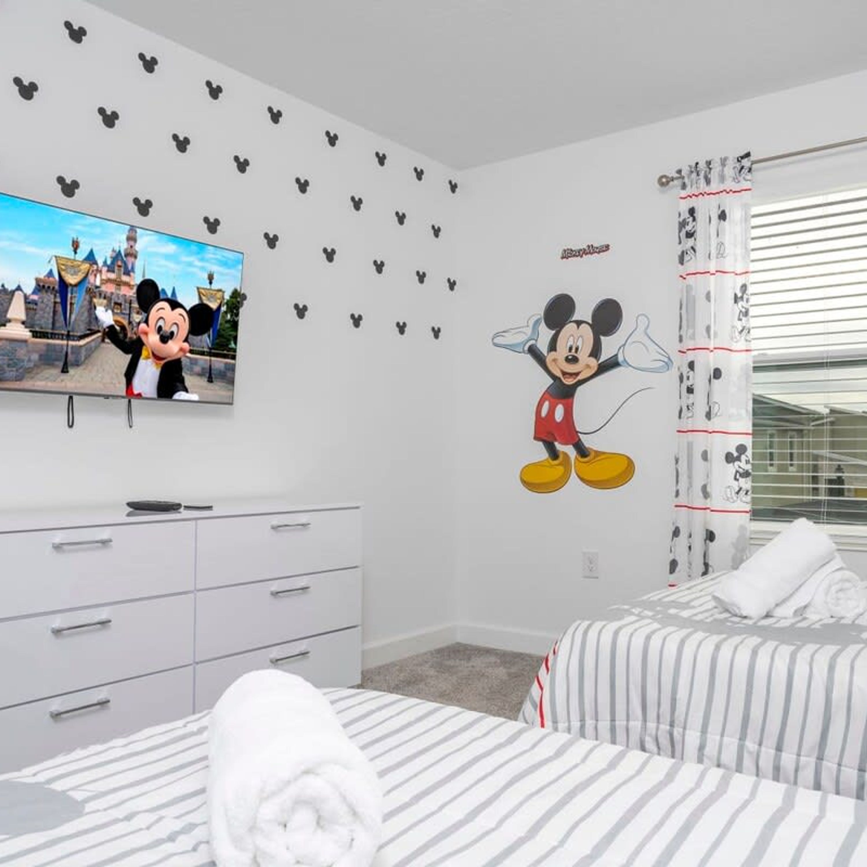 A hotel room with Mickey Mouse wall decals, a television, a bed with striped bedding, and a window with blinds.