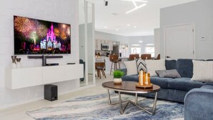 A modern living room with a flat-screen TV mounted on the wall, a blue sofa, a coffee table, and a kitchen in the background. - 150 Stunning 4BD Perfect for Your Vacation (Davenport)