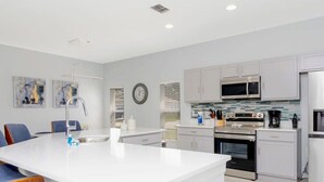 A modern kitchen with white cabinetry, a central island, stainless steel appliances, and a dining area with blue chairs. - 150 Stunning 4BD Perfect for Your Vacation (Davenport)