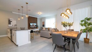 A modern living room with a dining area, kitchen, and a TV in the background. - 2681 Stunning 4BD Home Perfect For Your Family (Kissimmee)