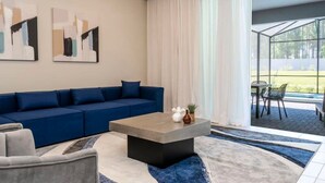 Ein modernes Wohnzimmer mit einem blauen Sofa, einem grauen Sessel und einem Couchtisch. An den Wänden hängen abstrakte Gemälde und ein großes Fenster mit Vorhängen.