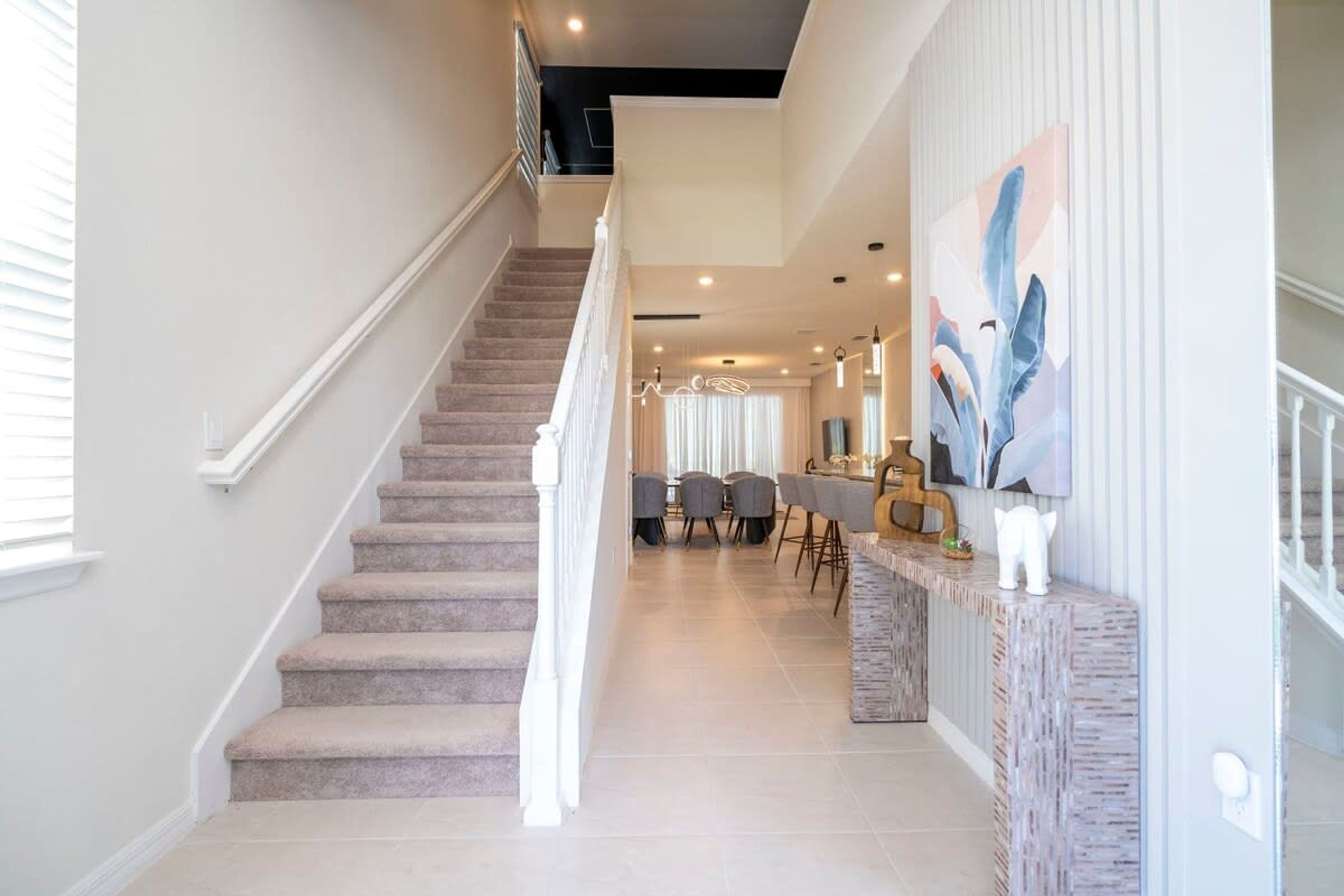 Una escalera conduce a un comedor con una lámpara de araña moderna y un cuadro abstracto de gran tamaño en la pared.