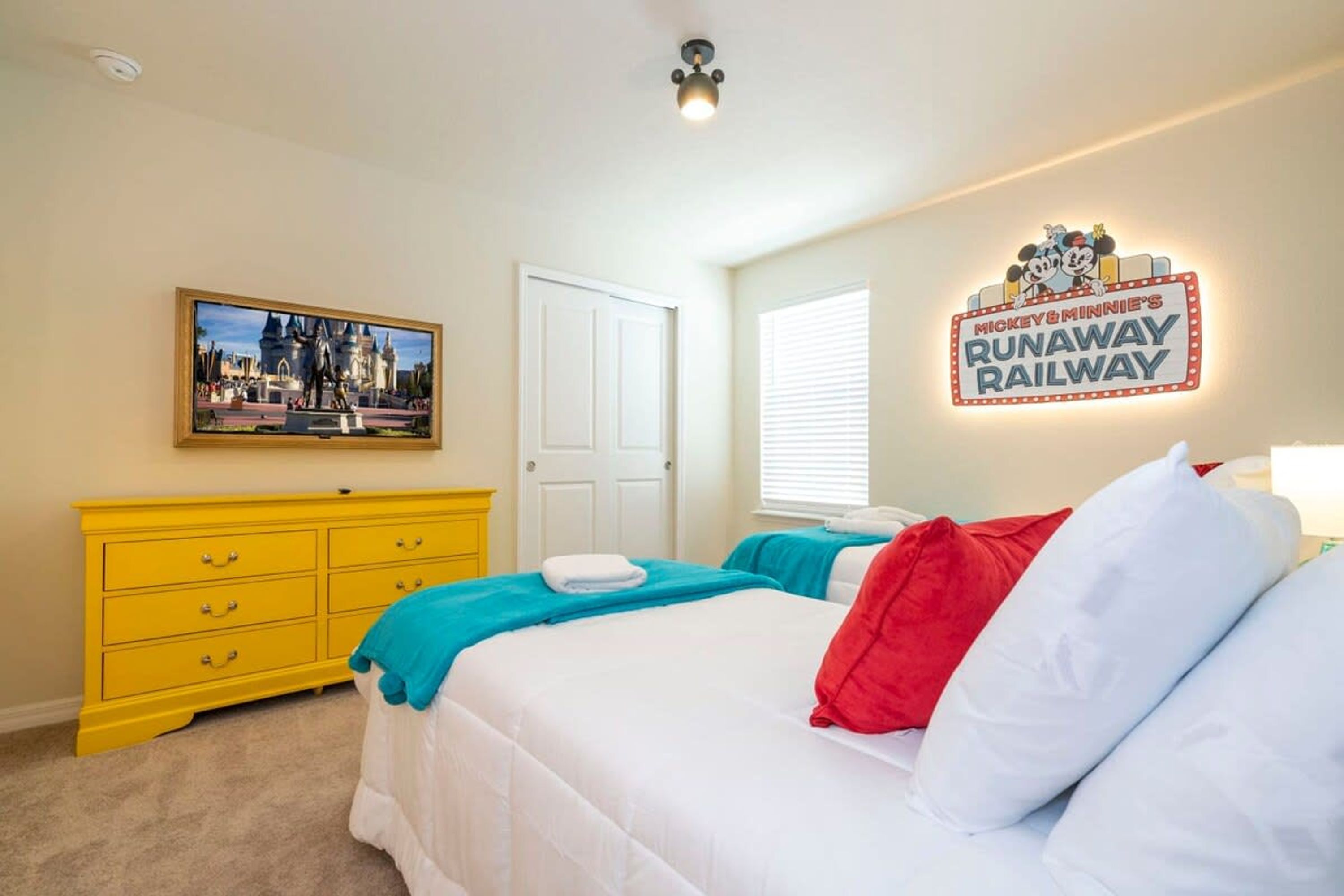 A hotel room with a bed, a yellow dresser, a Mickey & Minnie sign, and a framed picture on the wall.