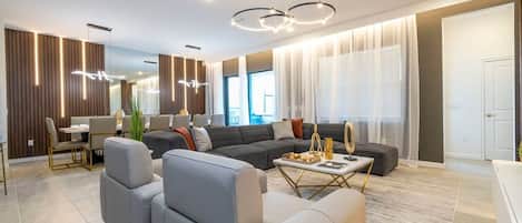 A modern living room with a sectional sofa, a dining area with a table and chairs, and a large window with sheer curtains.