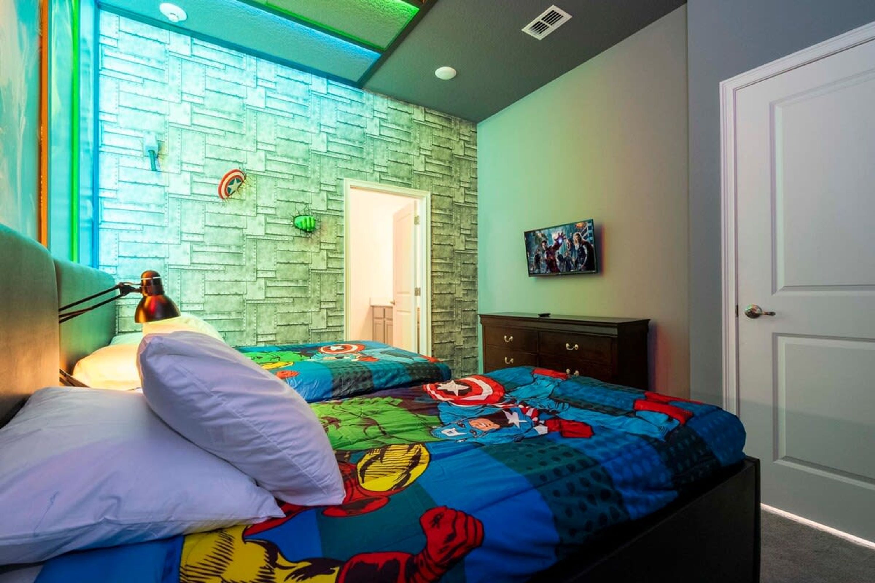 Quarto com parede em tijolos, cama com roupa de cama temática de super-heróis, abajur e porta para outro cômodo.
