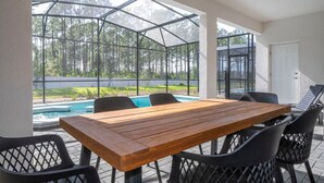 Eine überdachte Terrasse mit einem Holztisch und schwarzen Stühlen, mit Blick auf einen Swimmingpool und einen bewaldeten Bereich.