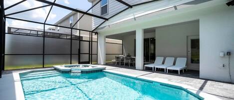 A swimming pool with a hot tub, surrounded by a covered patio area with lounge chairs and a table.