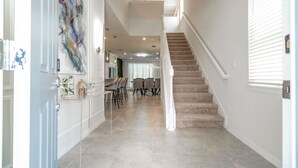 A spacious hallway with a staircase leading to an upper floor, a dining area with chairs and tables, and a decorative wall piece.