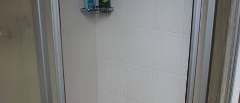 A small, enclosed shower area with a glass door, a white tiled wall, and a shelf with two toiletries.